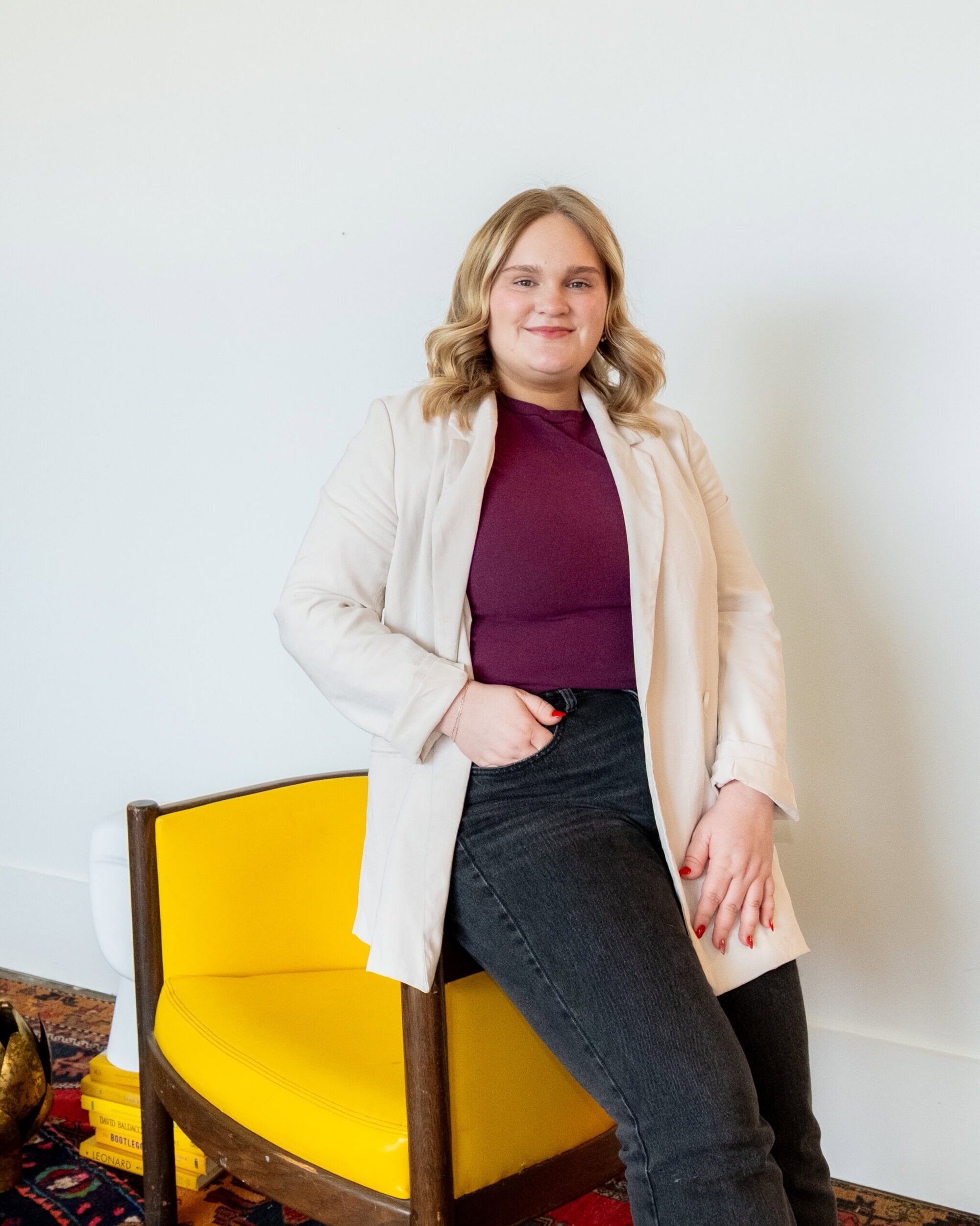 A person with blonde hair wearing a light blazer, maroon top, and dark jeans sits on the armrest of a yellow chair against a white wall.