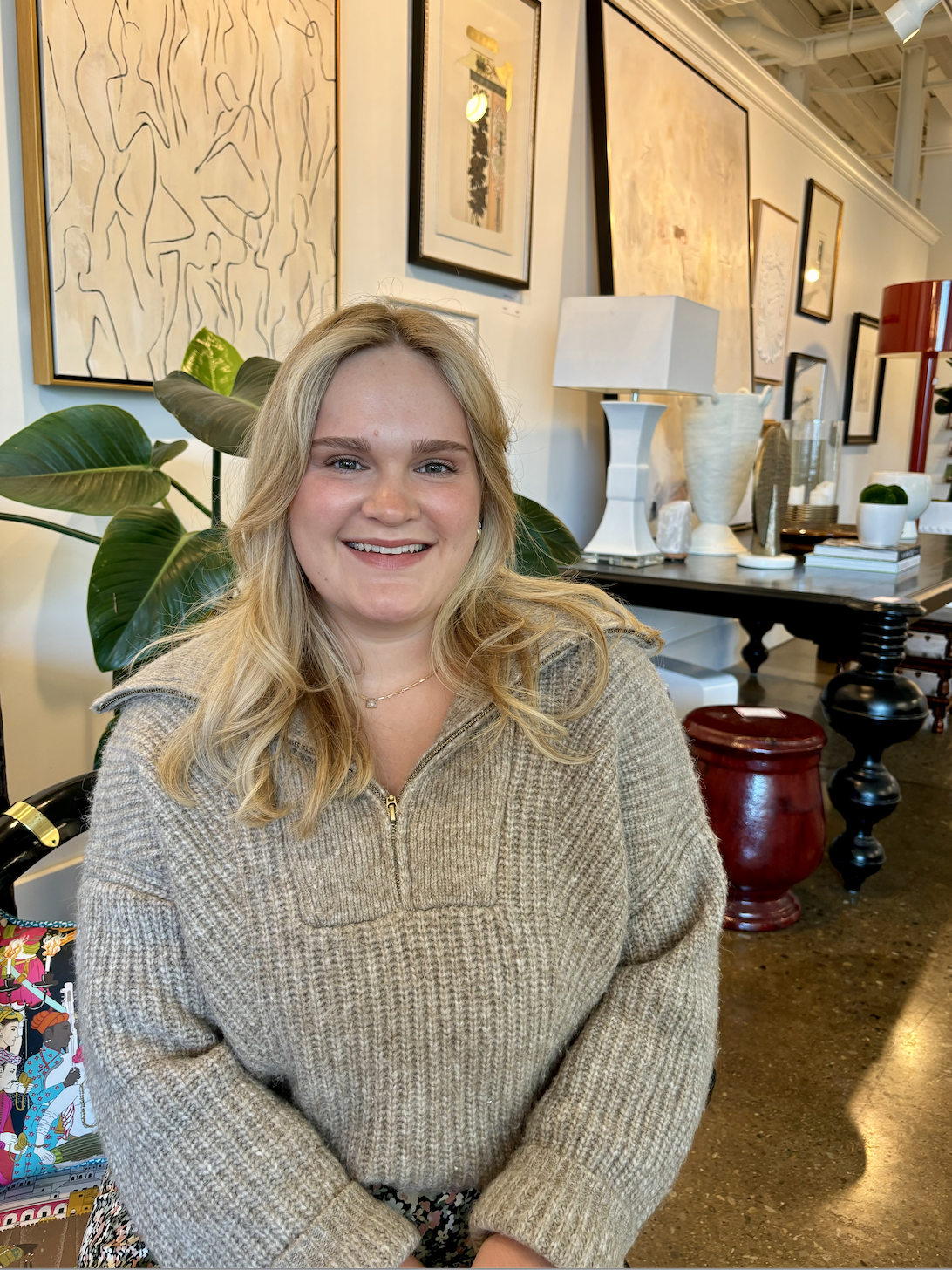 A woman with blonde hair and a beige sweater sits and smiles in a modern, art-filled room curated by Crimson Design Group, featuring plants and decorative objects in the background.