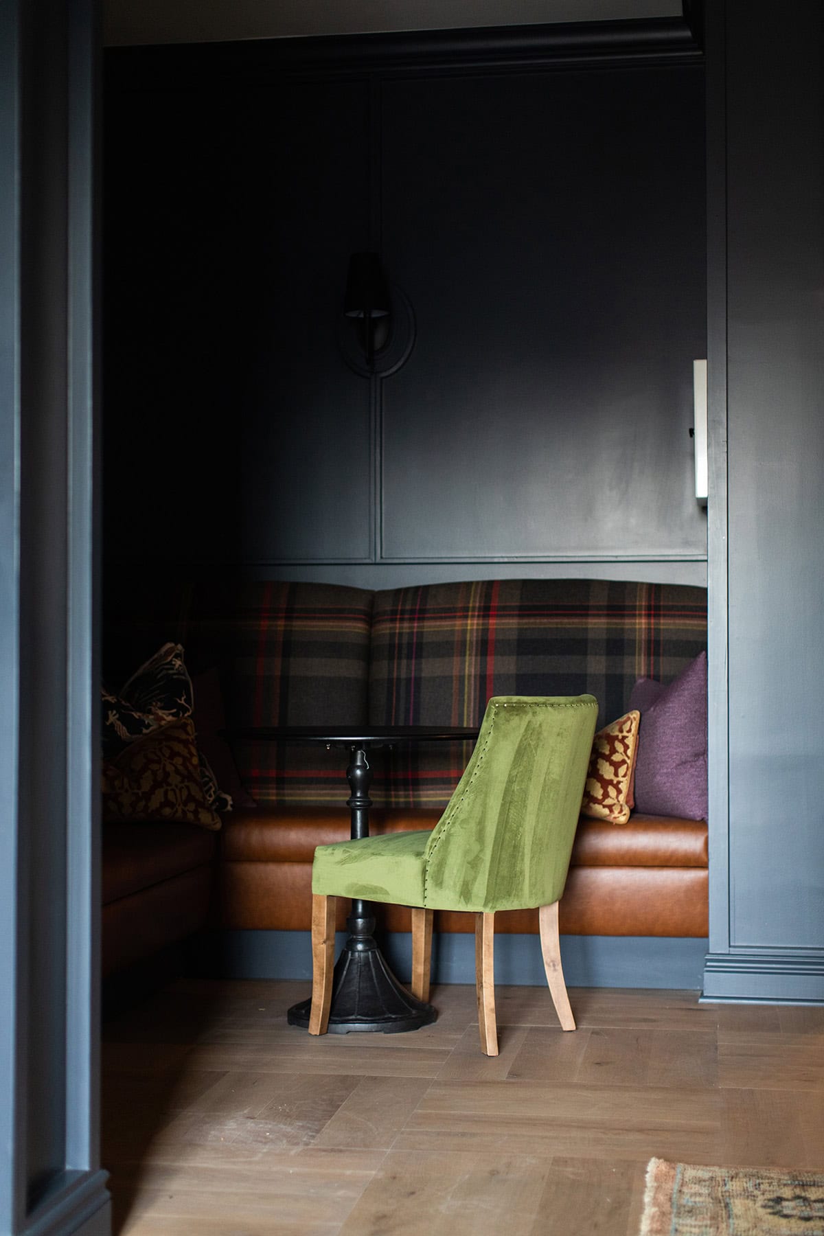 A small green chair is placed in front of a plaid upholstered bench in a dimly lit room with dark blue walls and wooden floor.