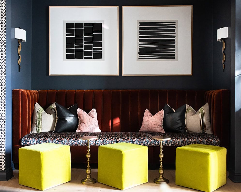 A modern seating area with a rust-colored velvet banquette, patterned pillows, three yellow cube ottomans, small brass tables, and abstract black-and-white artwork on a dark wall.