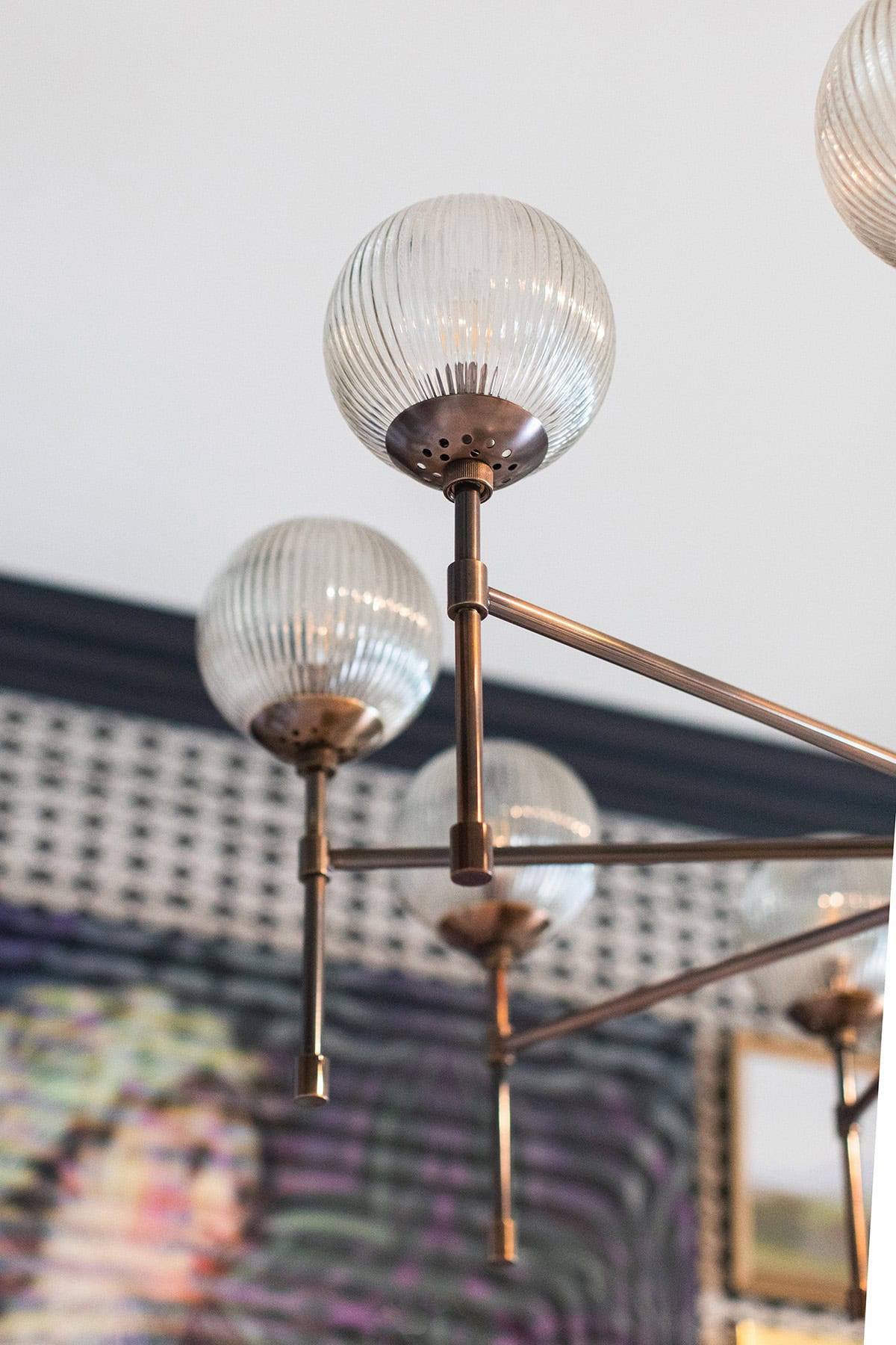 Close-up view of a modern chandelier with ribbed glass globes and metal arms, set against a patterned wall and framed artwork in the background.