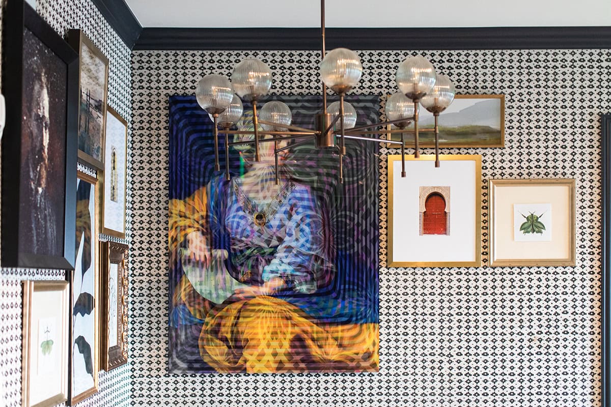 A chandelier hangs from the ceiling in a room with patterned wallpaper and various framed artworks, including a large colorful portrait and smaller landscape and insect-themed pieces.