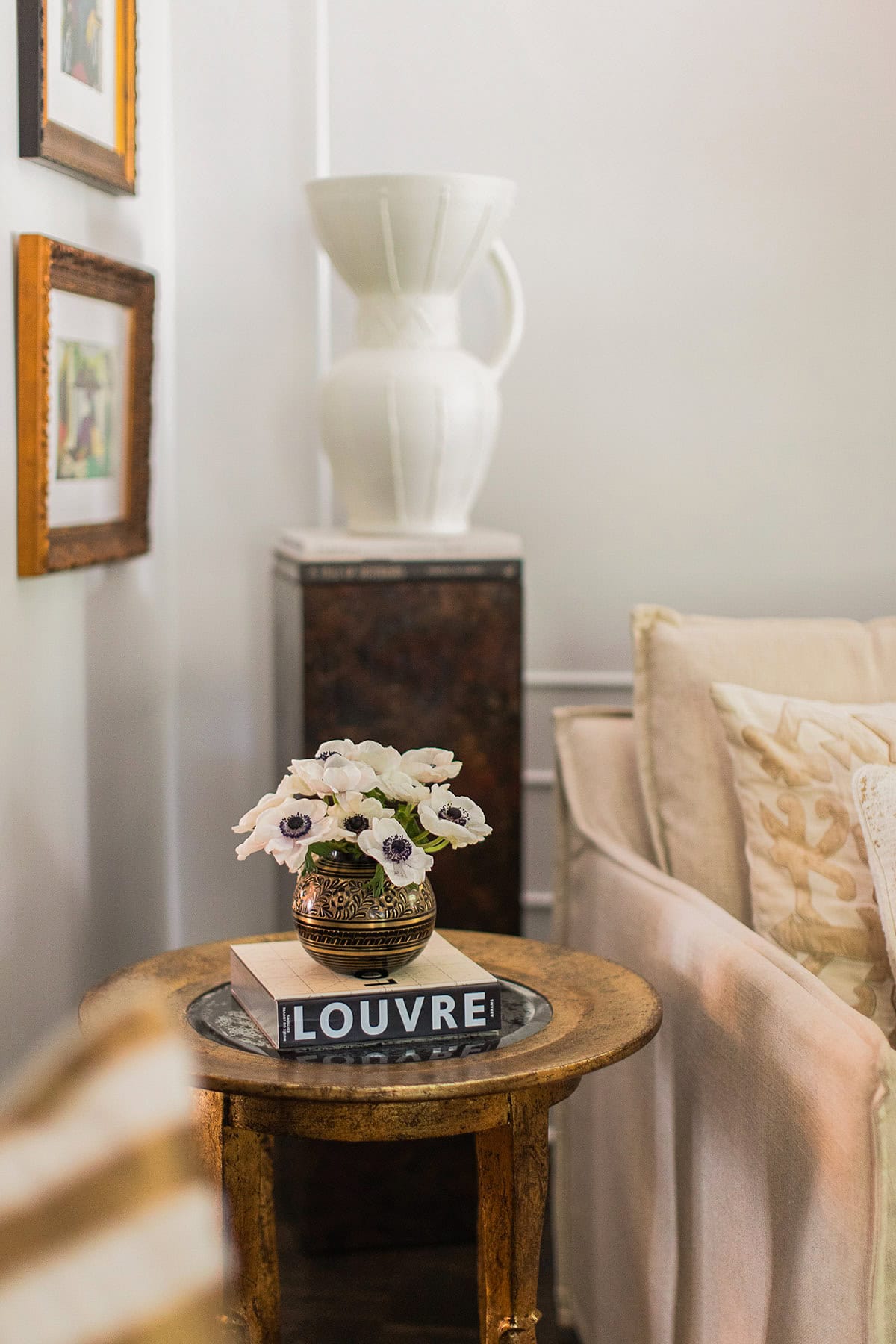 A small round table with a vase of white flowers and a book titled "LOUVRE" sits beside a beige sofa; a white ceramic jug is displayed on a pedestal in the background.