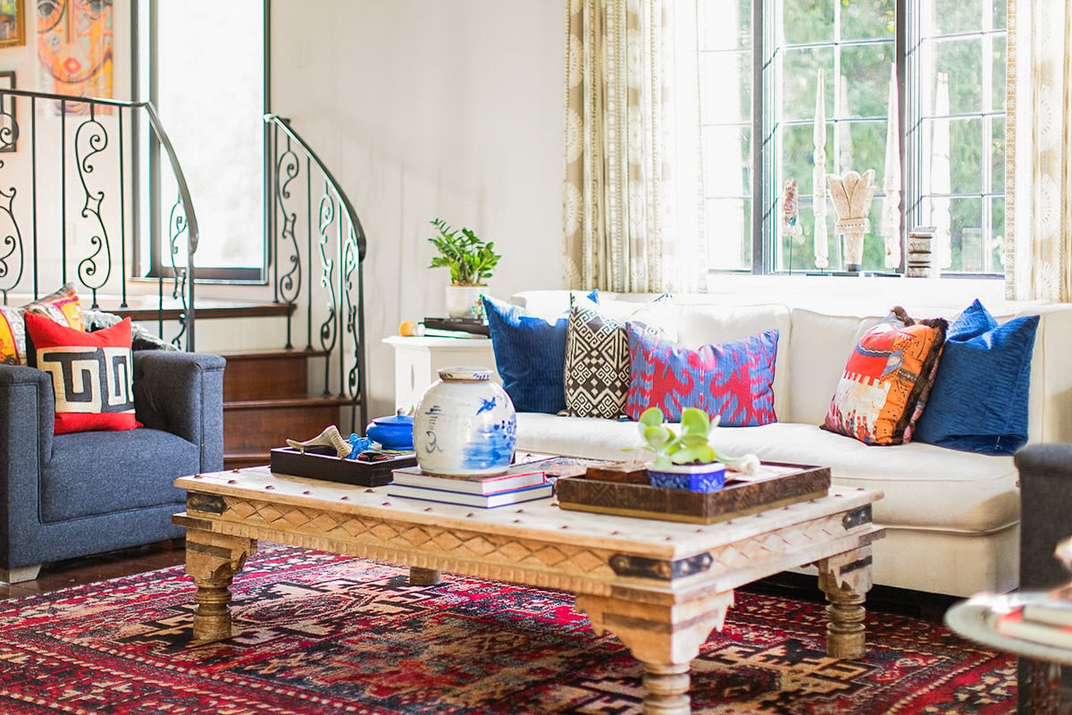 A bright living room with a white sofa, colorful patterned pillows, a wooden coffee table, a red patterned rug, and armchairs, near a staircase with a decorative railing.