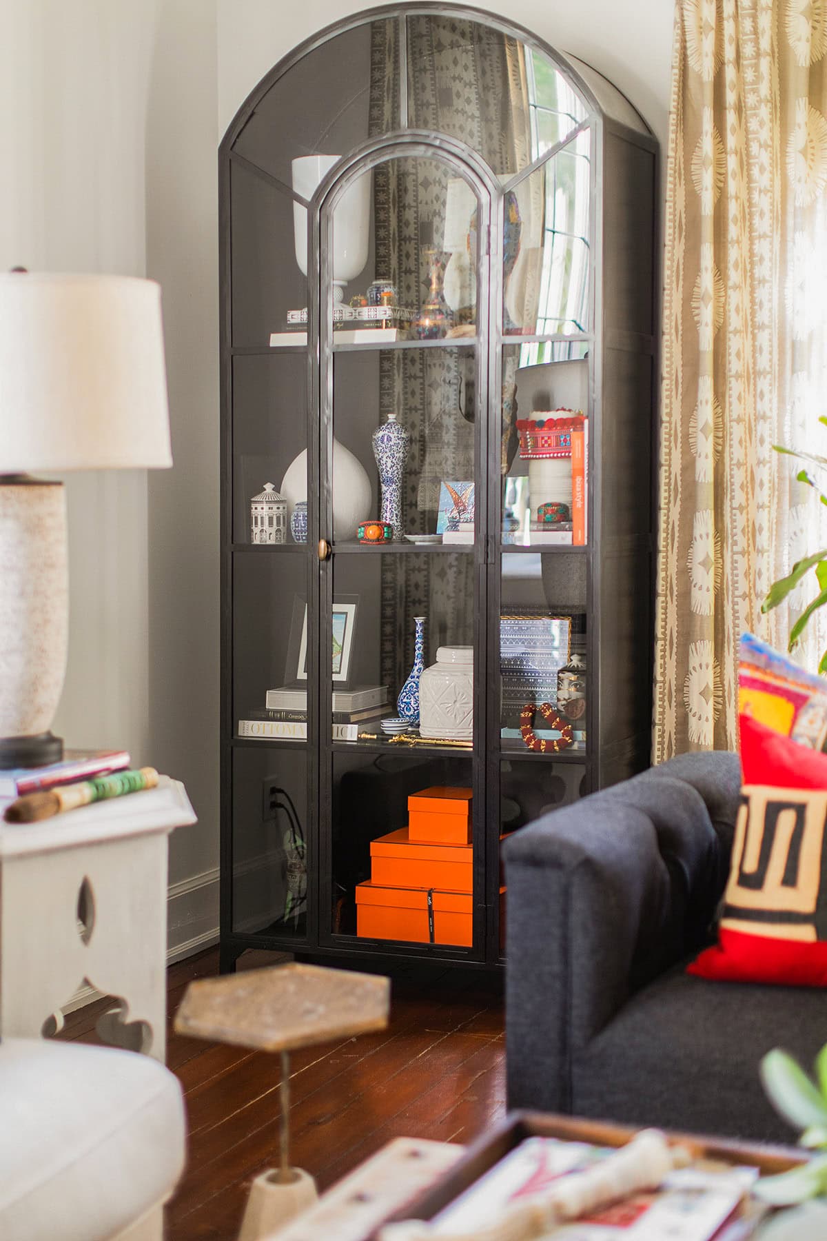 A black glass-front cabinet displays decorative objects, books, and orange boxes in the corner of a living room with a sofa, side table, lamp, and patterned curtains.