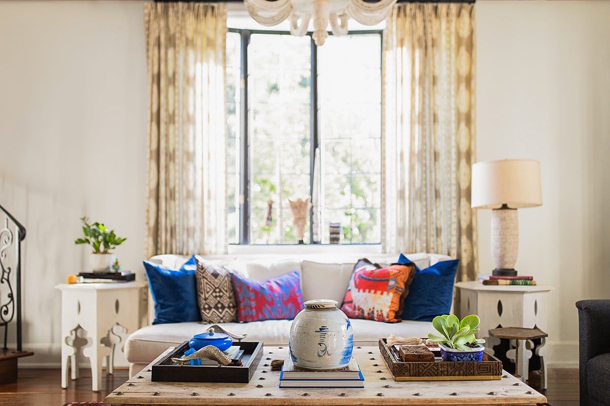 Bright living room with a white sofa, colorful pillows, a wooden coffee table with decor items, side tables, a lamp, and large window with sheer curtains.