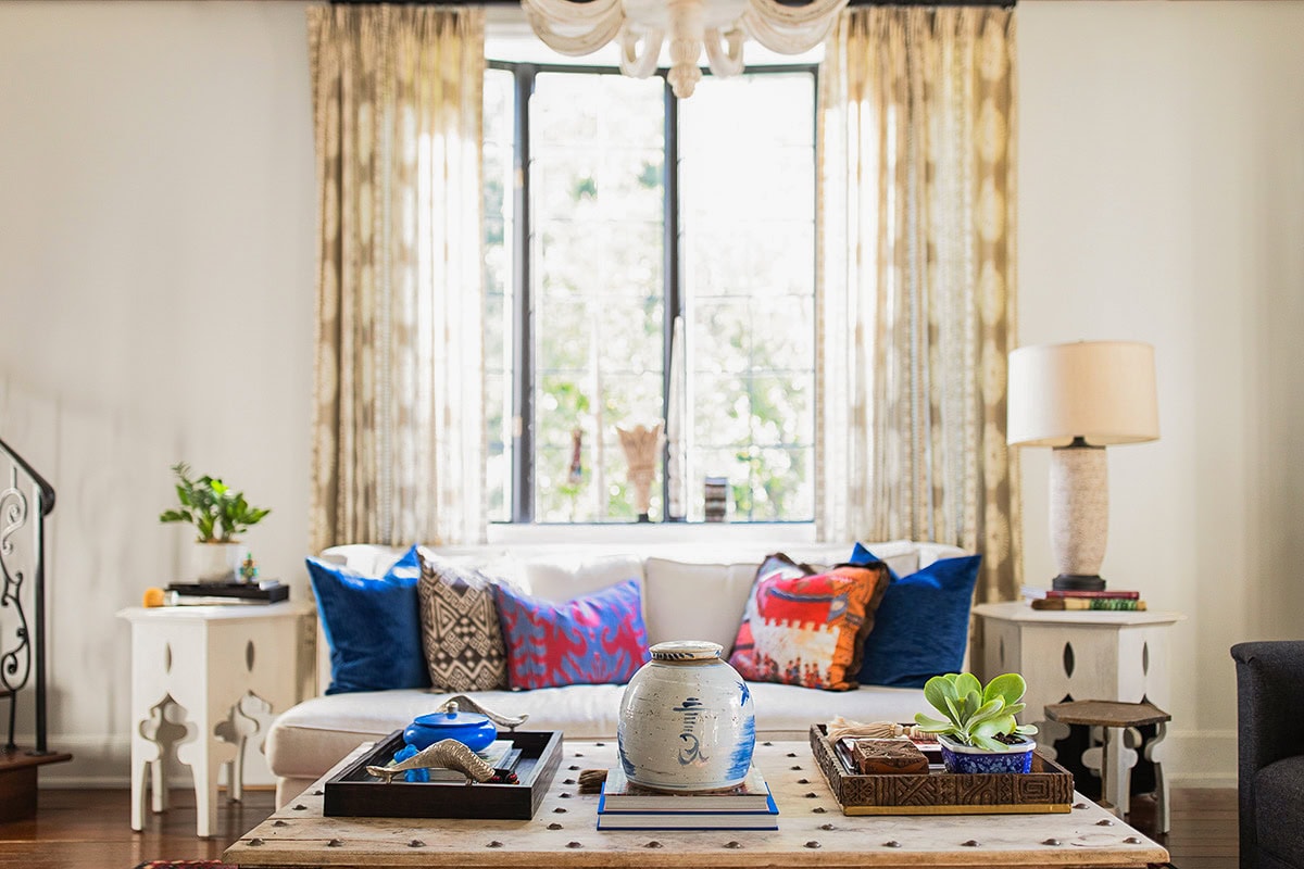 A living room with a white sofa, colorful pillows, a wooden coffee table with decor, side tables with plants, a lamp, and a large window with sheer curtains.