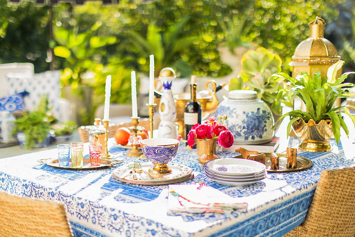 A table set outdoors with blue and white patterned tablecloth, assorted dishes, candles, glassware, vases, flowers, fruit, and decorative gold accents, surrounded by greenery.