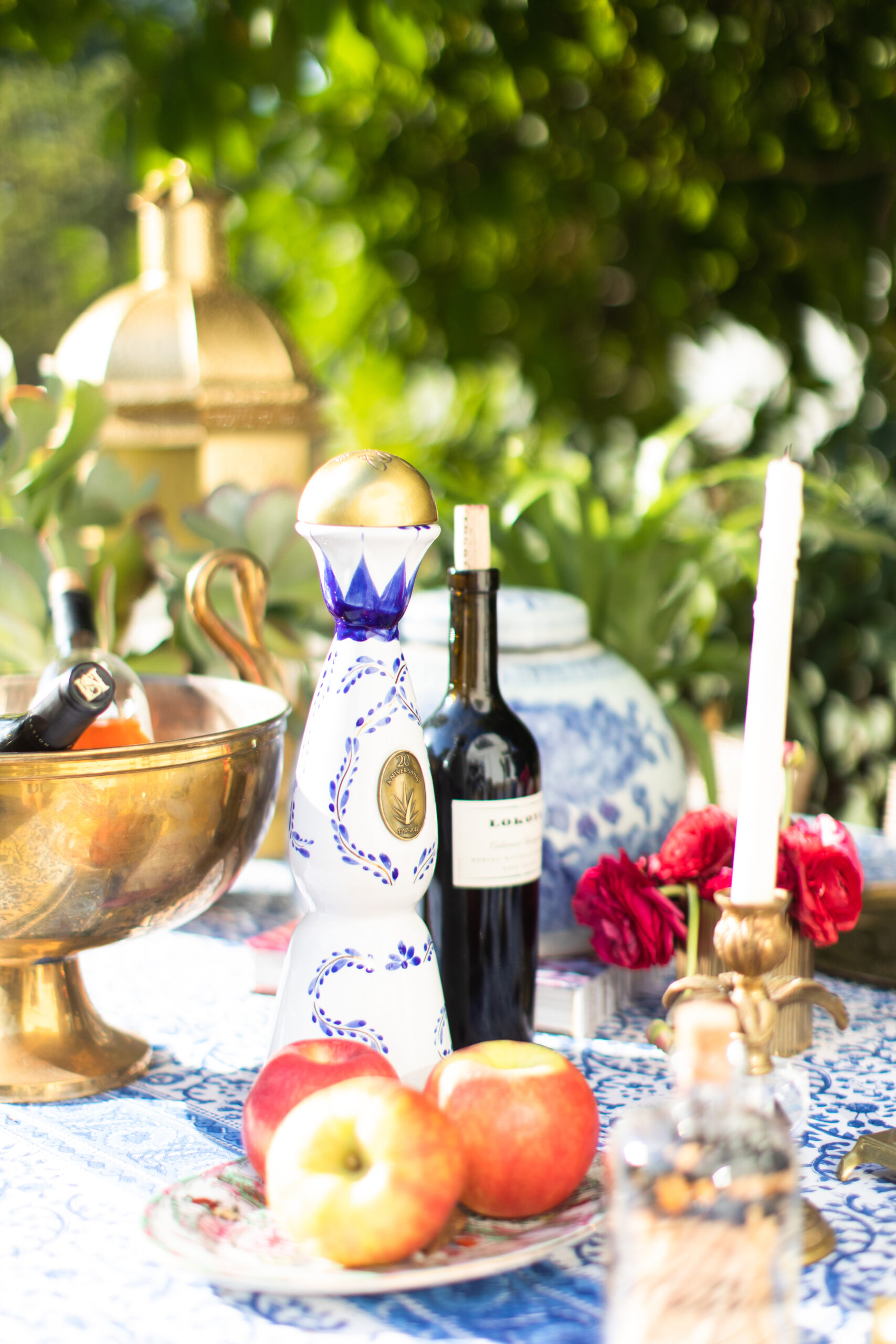 A bottle of tequila, a bottle of red wine, apples, a candle, and floral decor by Crimson Design Group are artfully arranged on a patterned table outdoors.