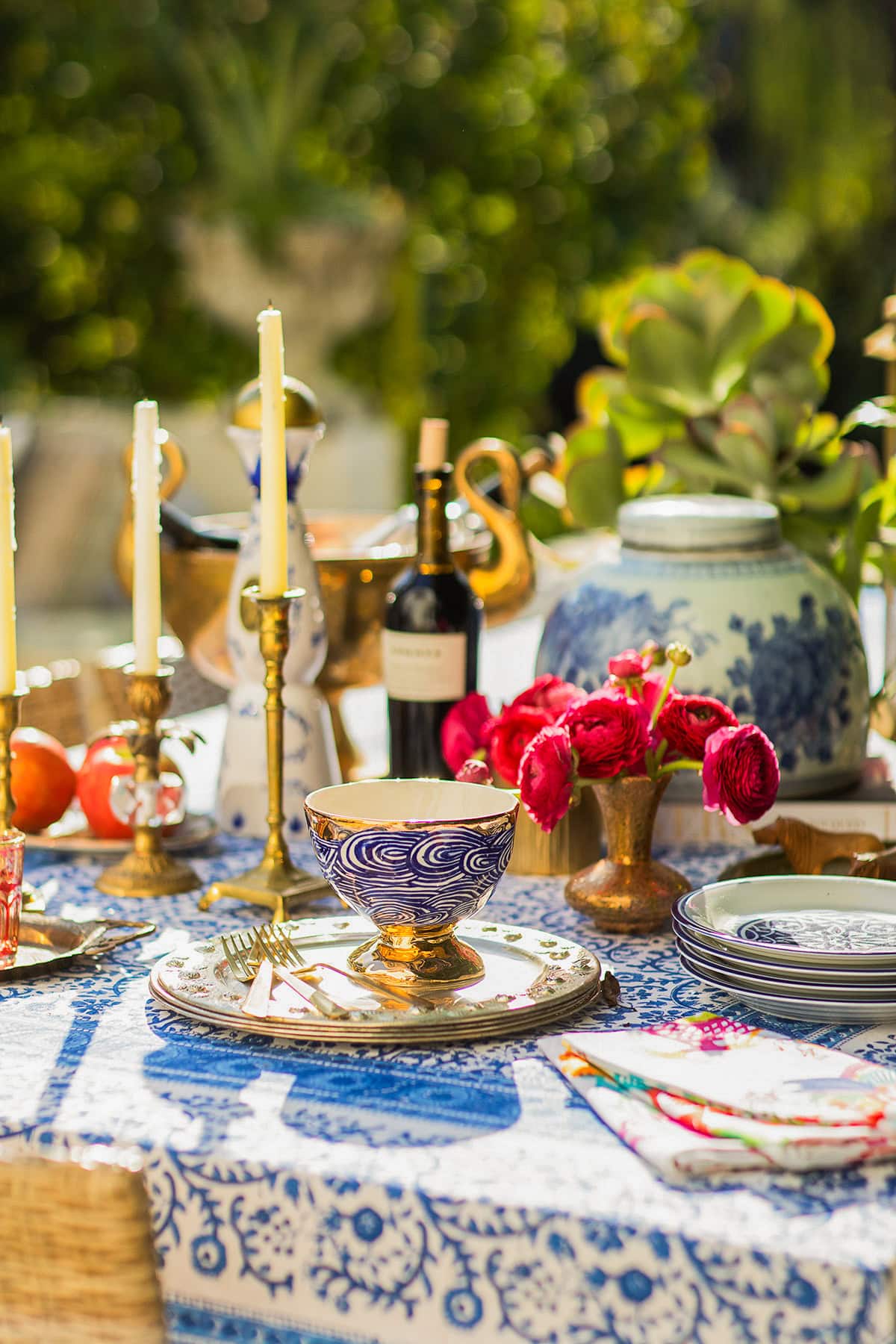 A colorful outdoor table setting with blue and white dishes, gold accents, red flowers, candles, a bottle of wine, and fruit on a patterned tablecloth.