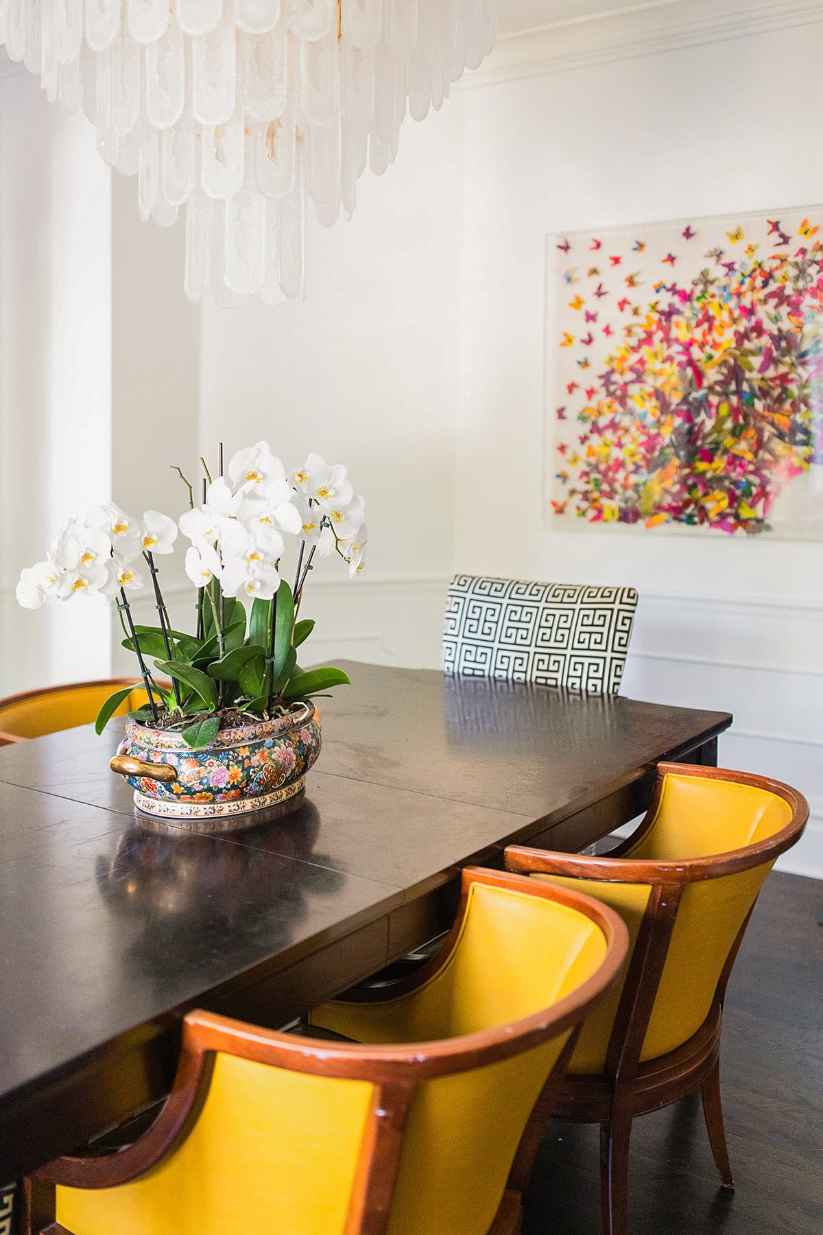 A dining room with a dark wood table, yellow chairs, a potted white orchid, a large chandelier, and colorful butterfly artwork on the wall.