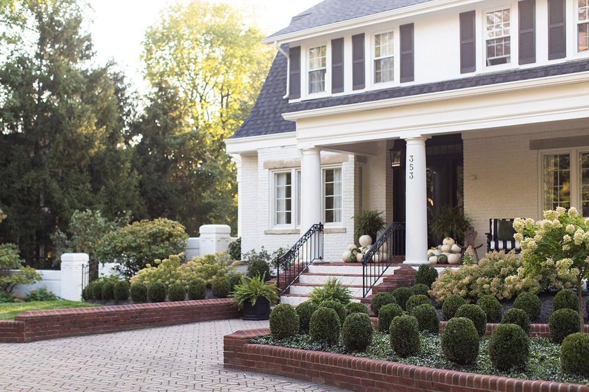 Wide shot of a large, well-maintained white colonial-style home with a black roof and shutters. A patterned brick paver driveway leads past brick-retained formal boxwood parterre gardens toward the front steps. Large white columns support the front porch, which is decorated with large ferns and potted plants, showing the house number 353. The entire scene is bathed in soft, golden hour sunlight.