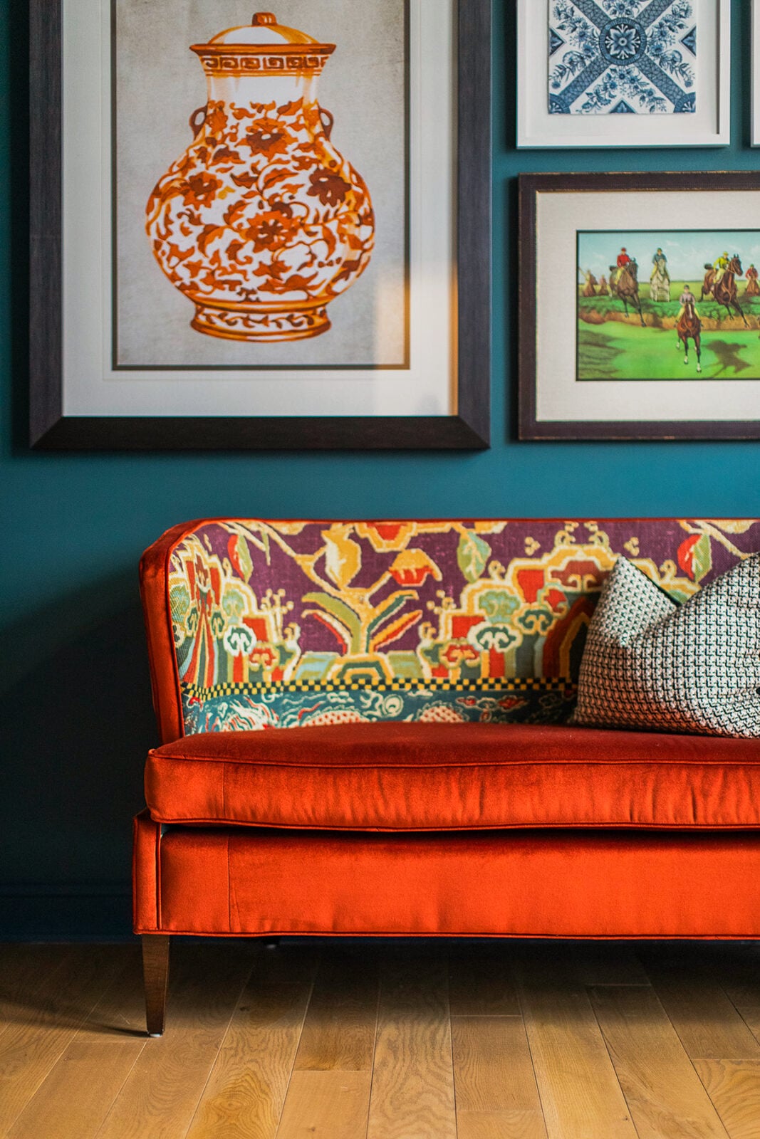 Bright orange velvet sofa with patterned backrest, two textured pillows, and framed artwork on a teal wall above a wooden floor.