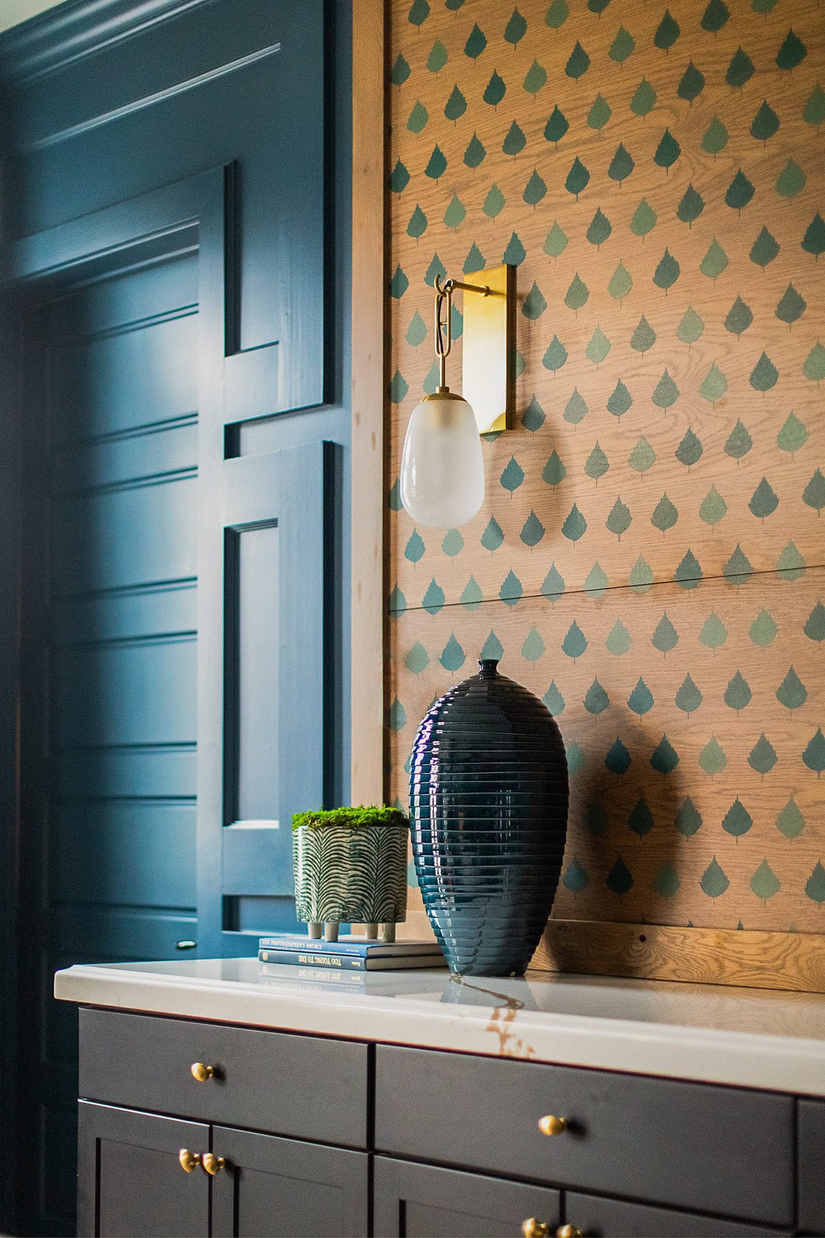 A modern interior features a navy cabinet with gold hardware, a white countertop, a blue vase, a small plant, patterned wallpaper, and a wall sconce with a frosted glass shade.