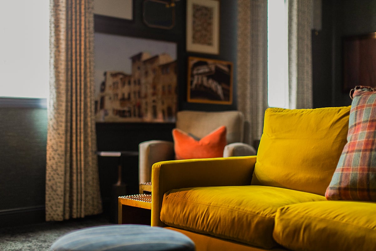 A yellow sofa with a plaid pillow sits in a living room, with a gray armchair and orange pillow in the background near windows with patterned curtains and wall art.