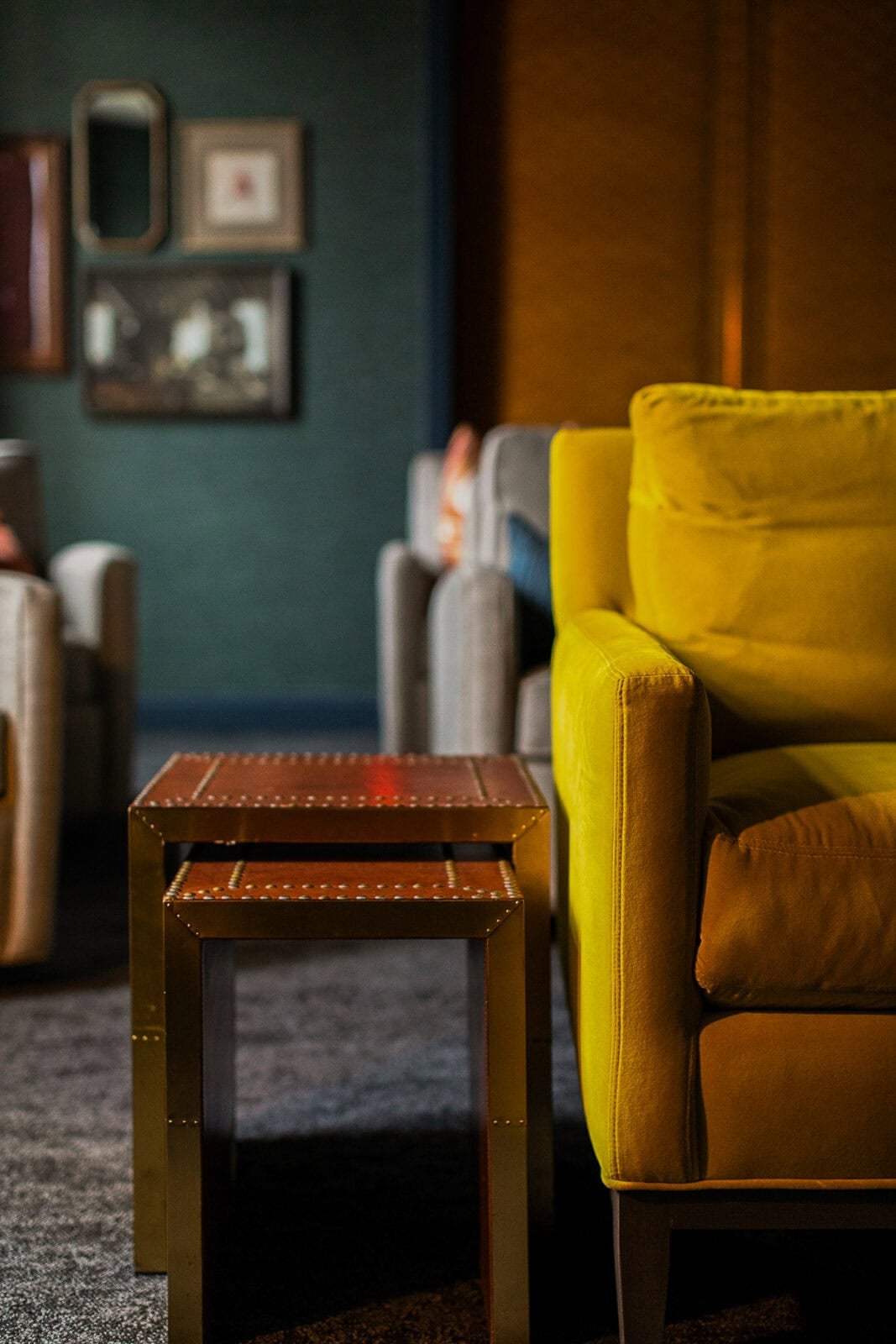 Yellow armchair next to two nesting tables with brass frames in a living room; framed art and another sofa are visible in the background.