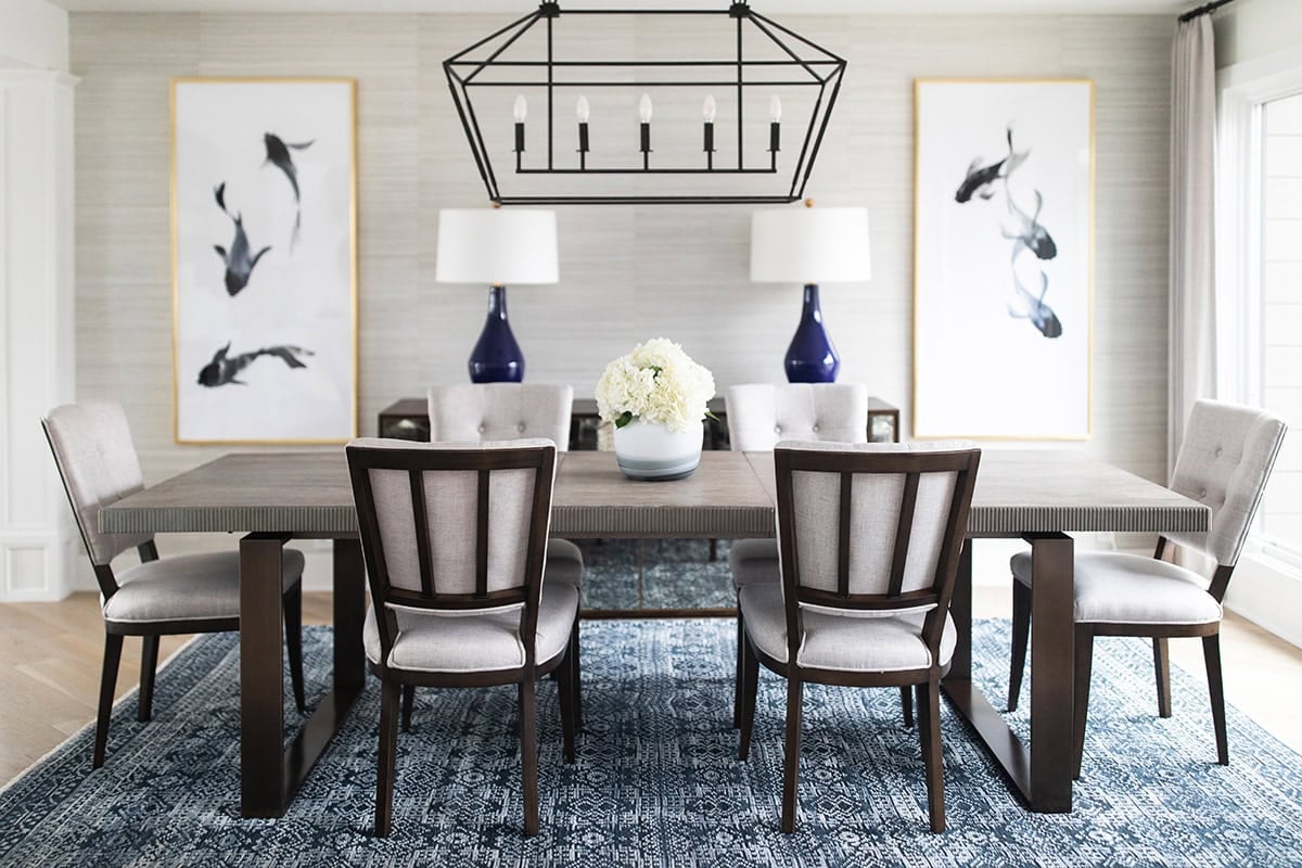 A modern dining room with a wooden table, six cushioned chairs, two blue lamps, floral centerpiece, abstract art, and a geometric chandelier over a blue patterned rug.