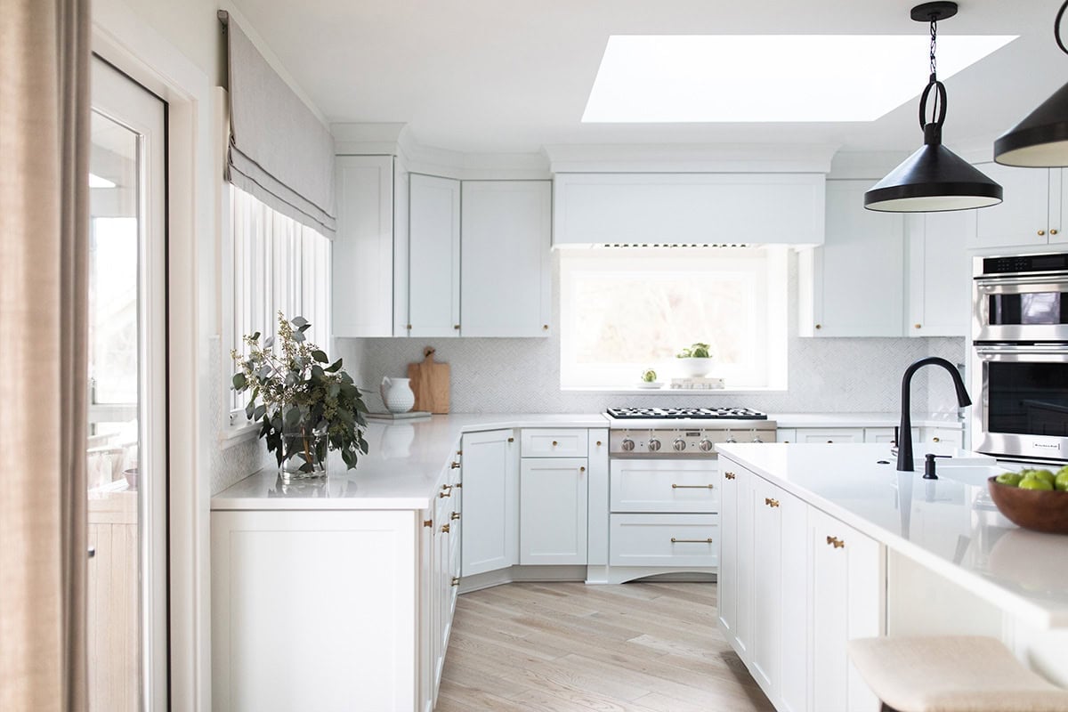 Bright, modern kitchen with white cabinets, a central island, black pendant lights, stainless steel appliances, and a window above the stove letting in natural light.