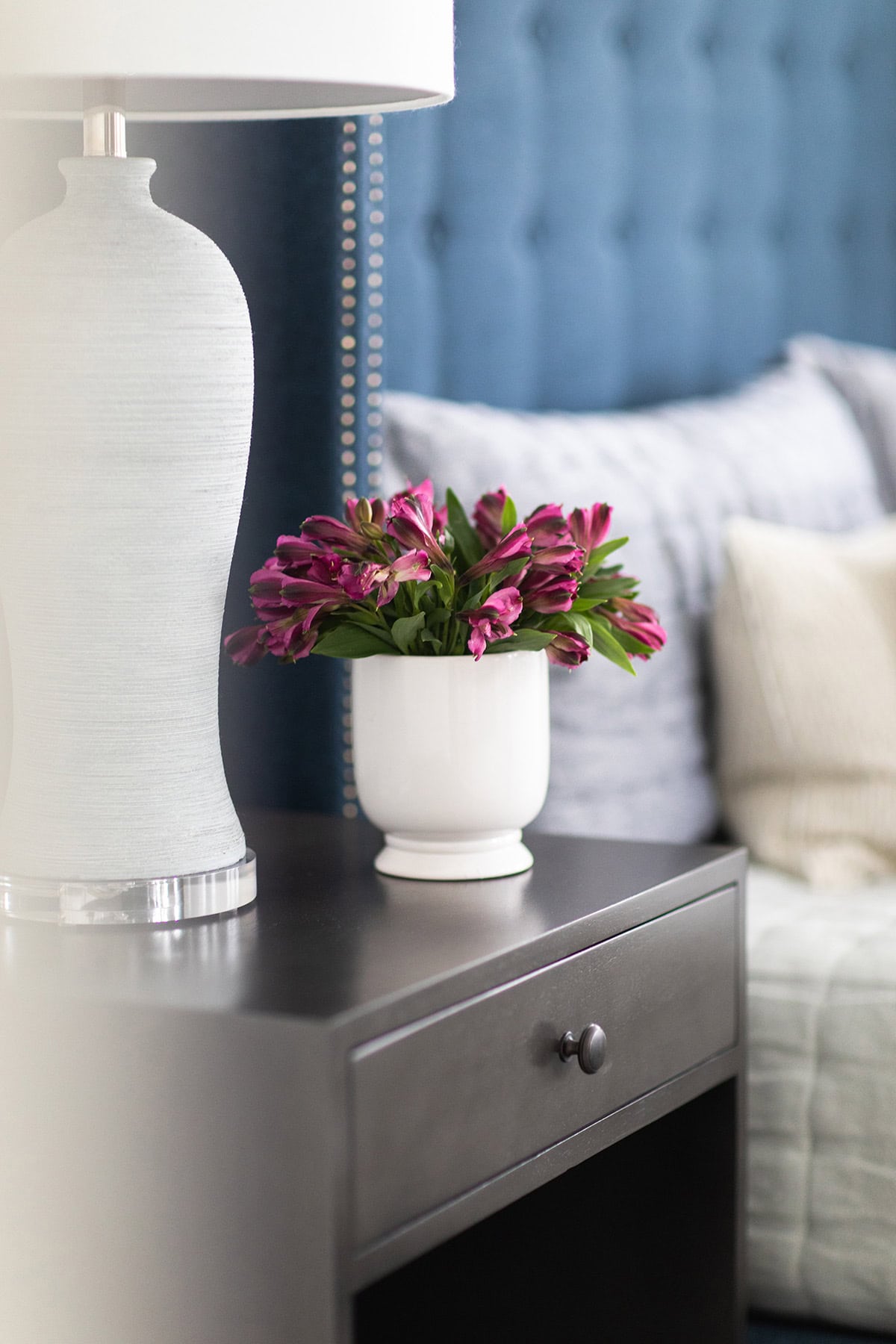 A white lamp and a white vase with purple flowers sit on a black nightstand next to a bed with blue and white bedding.