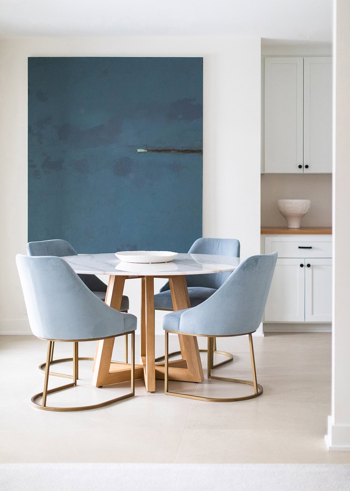 A modern dining area with a round marble table, four blue velvet chairs, a large blue abstract painting, and white cabinetry in the background.