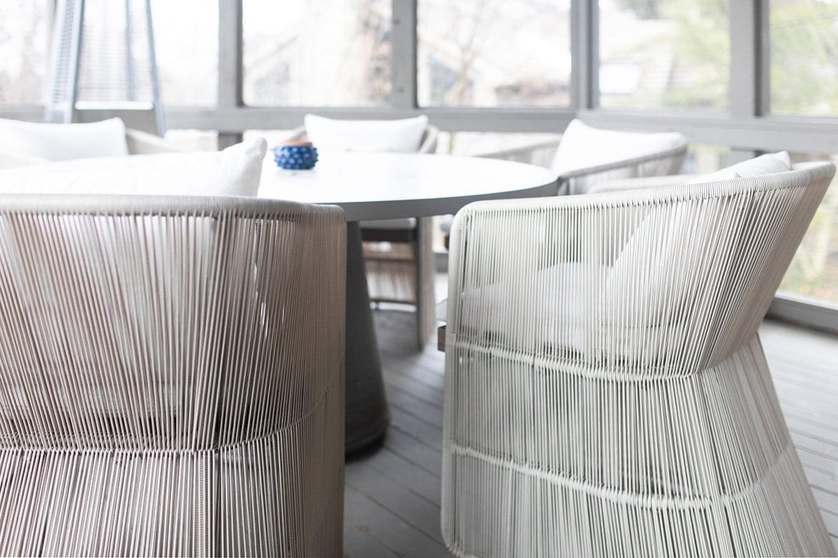 White string chairs with cushions surround a round white table on a light gray screened porch with natural light coming through the windows.
