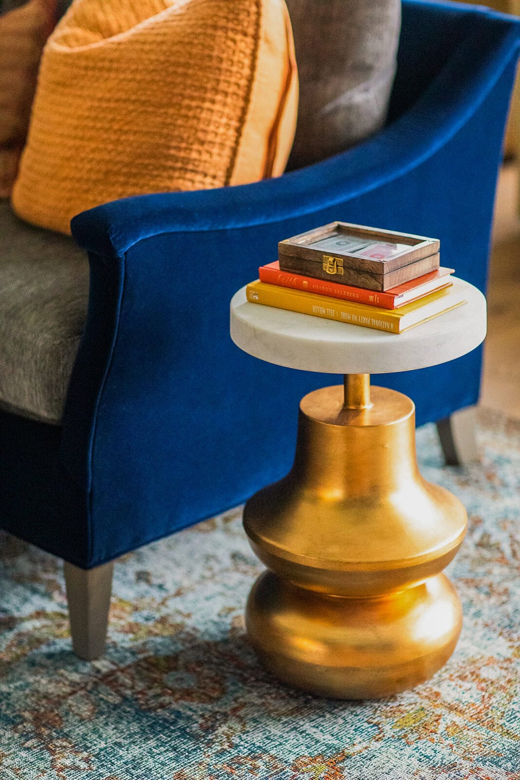 A small round side table with a gold base and white top holds a wooden box and stacked books beside a blue velvet sofa with orange and gray cushions on a patterned rug.