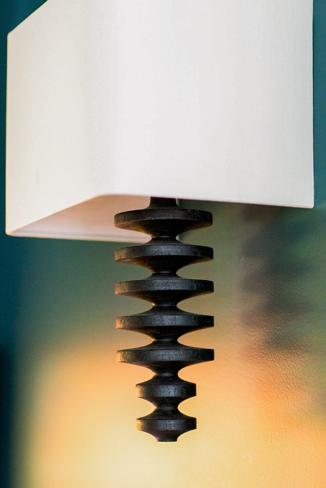 A wall-mounted lamp with a white rectangular shade and a textured, black, vertical decorative element beneath it, casting light on the wall.