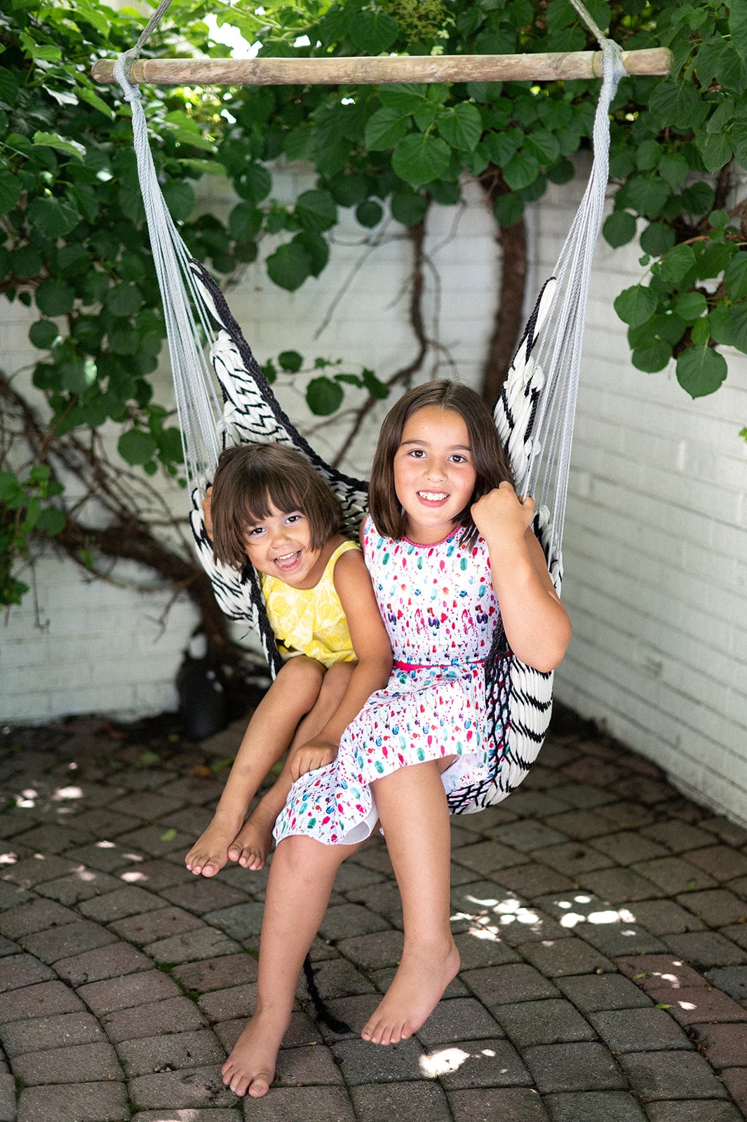 Two young children sit together on a hammock chair outdoors, both smiling at the camera, surrounded by greenery and a white brick wall—a charming scene inspired by Crimson Design Group style.