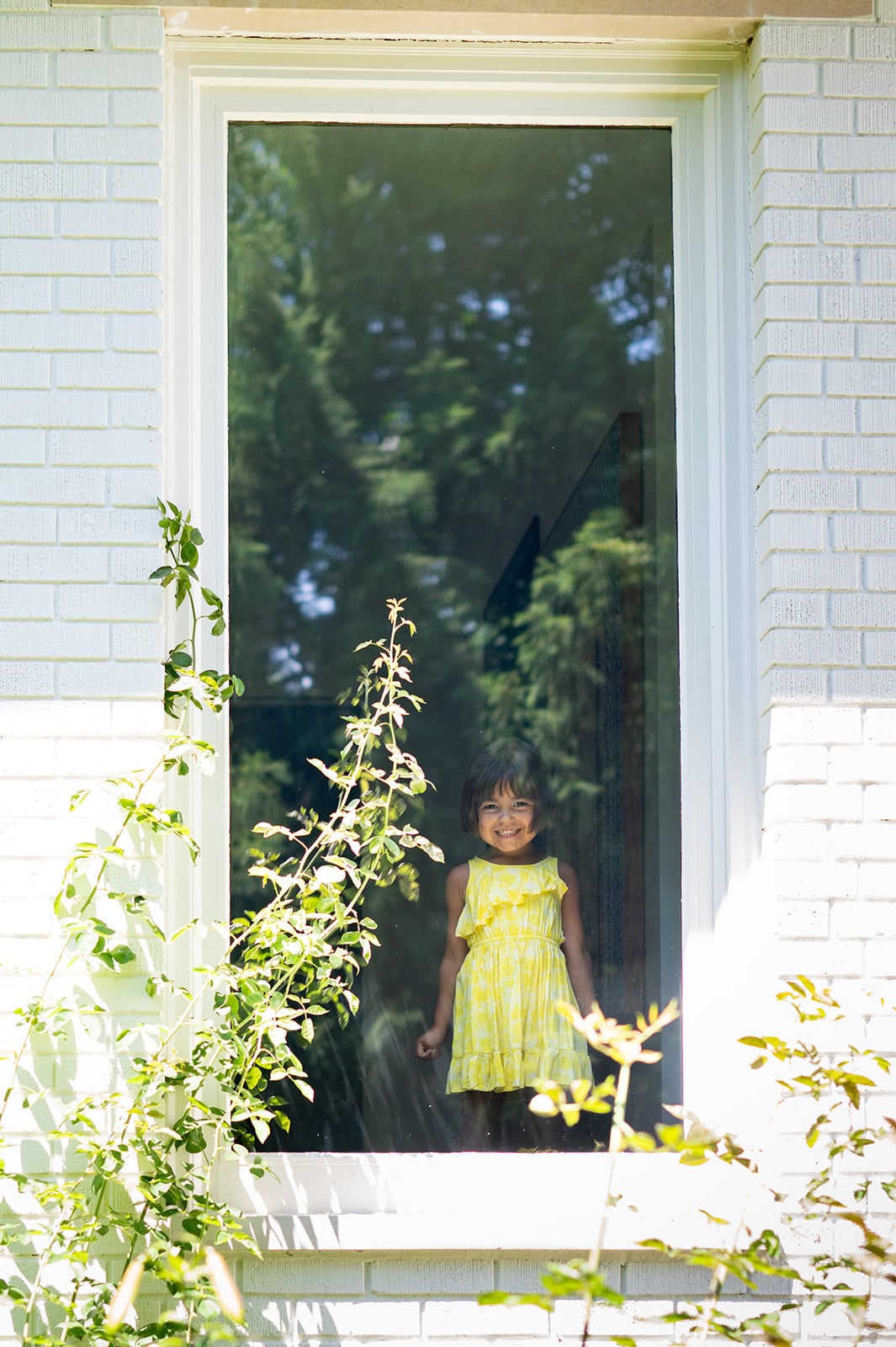 A young girl in a yellow dress stands and smiles behind a large window, with greenery and sunlight visible outside.