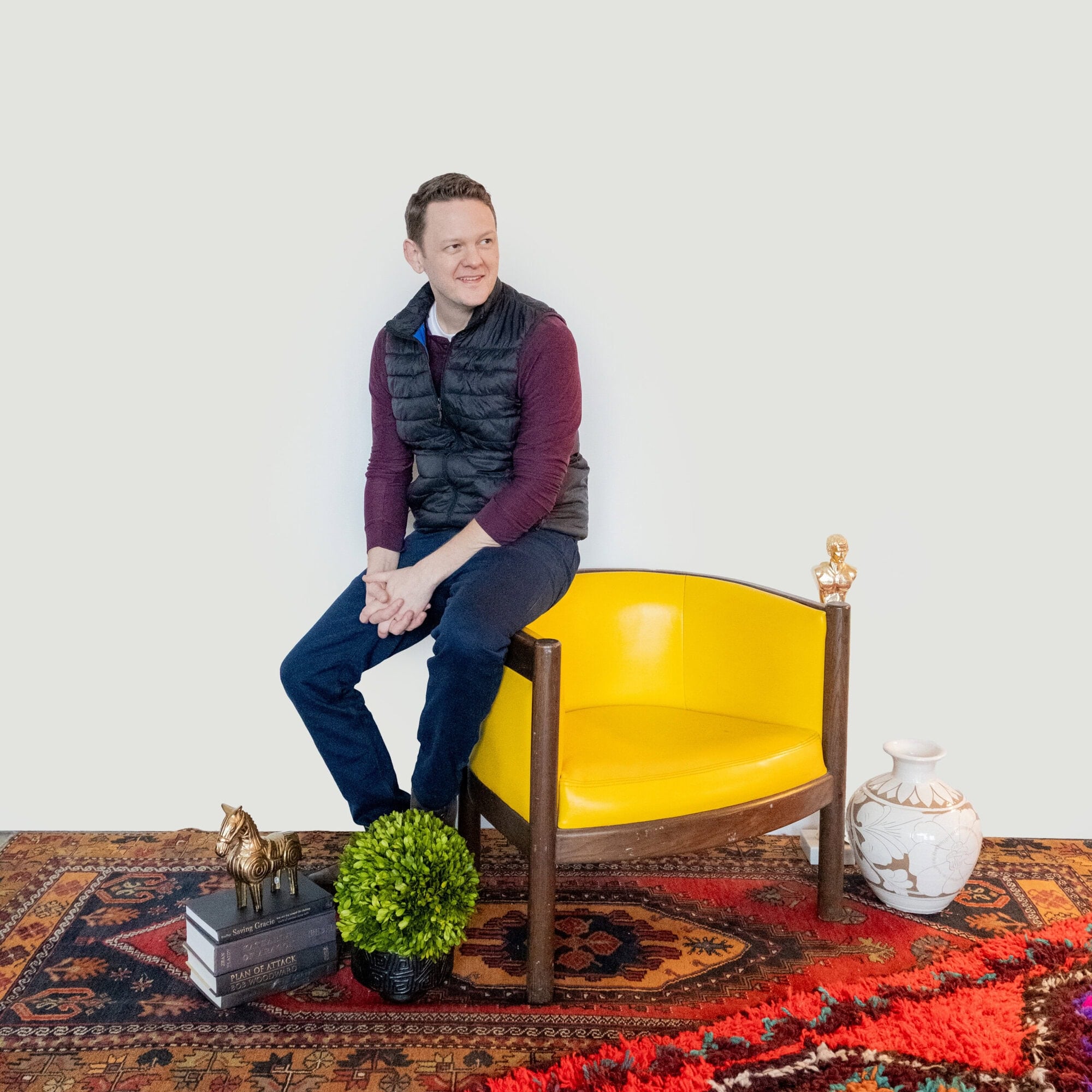 A man sitting on the arm of a yellow armchair on a patterned rug.