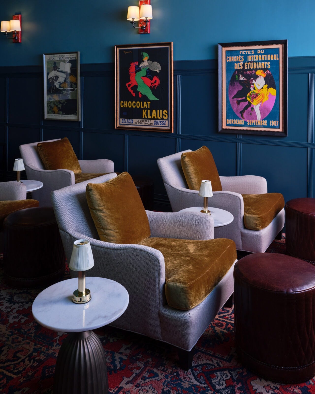 A lounge with velvet-cushioned armchairs, round side tables, wall lamps, and vintage posters on blue-paneled walls.