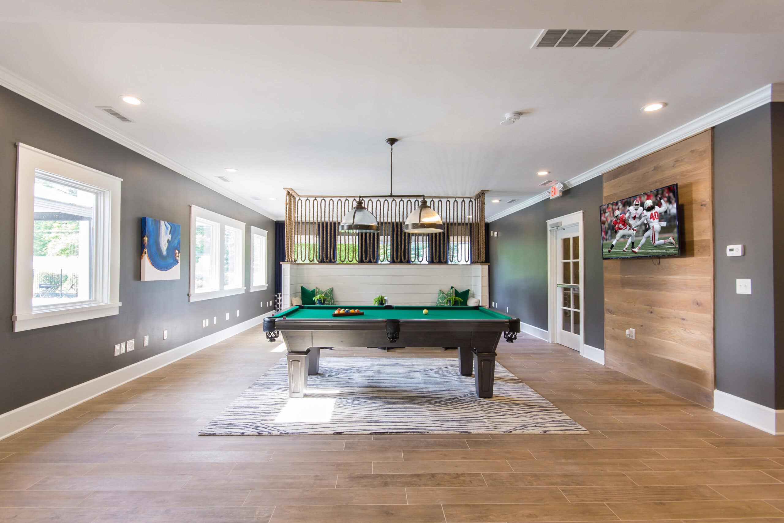 A modern game room with a pool table in the center, a TV on the wood-paneled wall showing a football game, and large windows providing natural light.