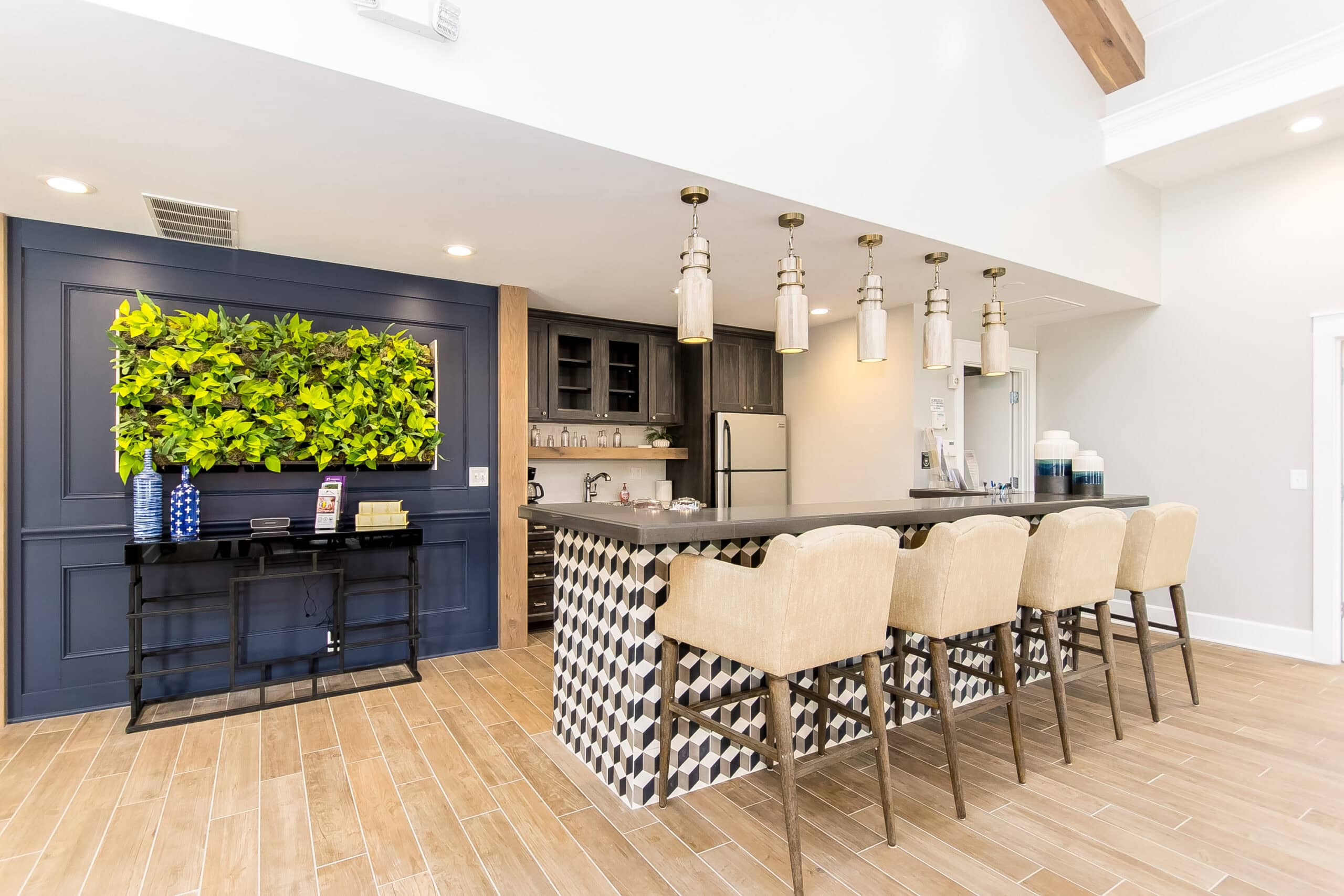Modern kitchen area with a patterned island, four beige barstools, hanging pendant lights, a vertical plant display, wood flooring, and a table with water dispensers.