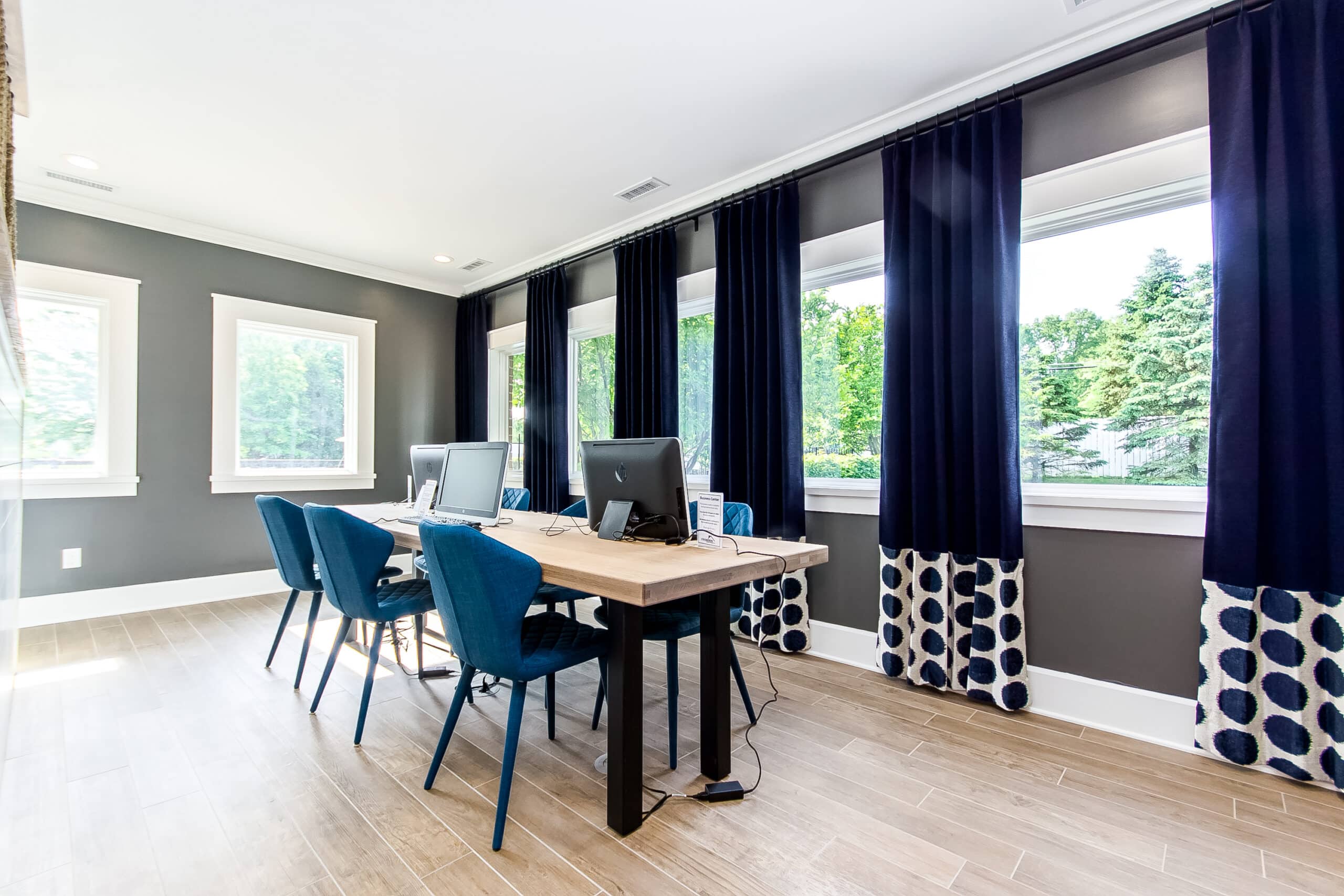 Modern office space with three blue chairs, a wooden desk with two computers, and large windows with dark blue curtains overlooking greenery.