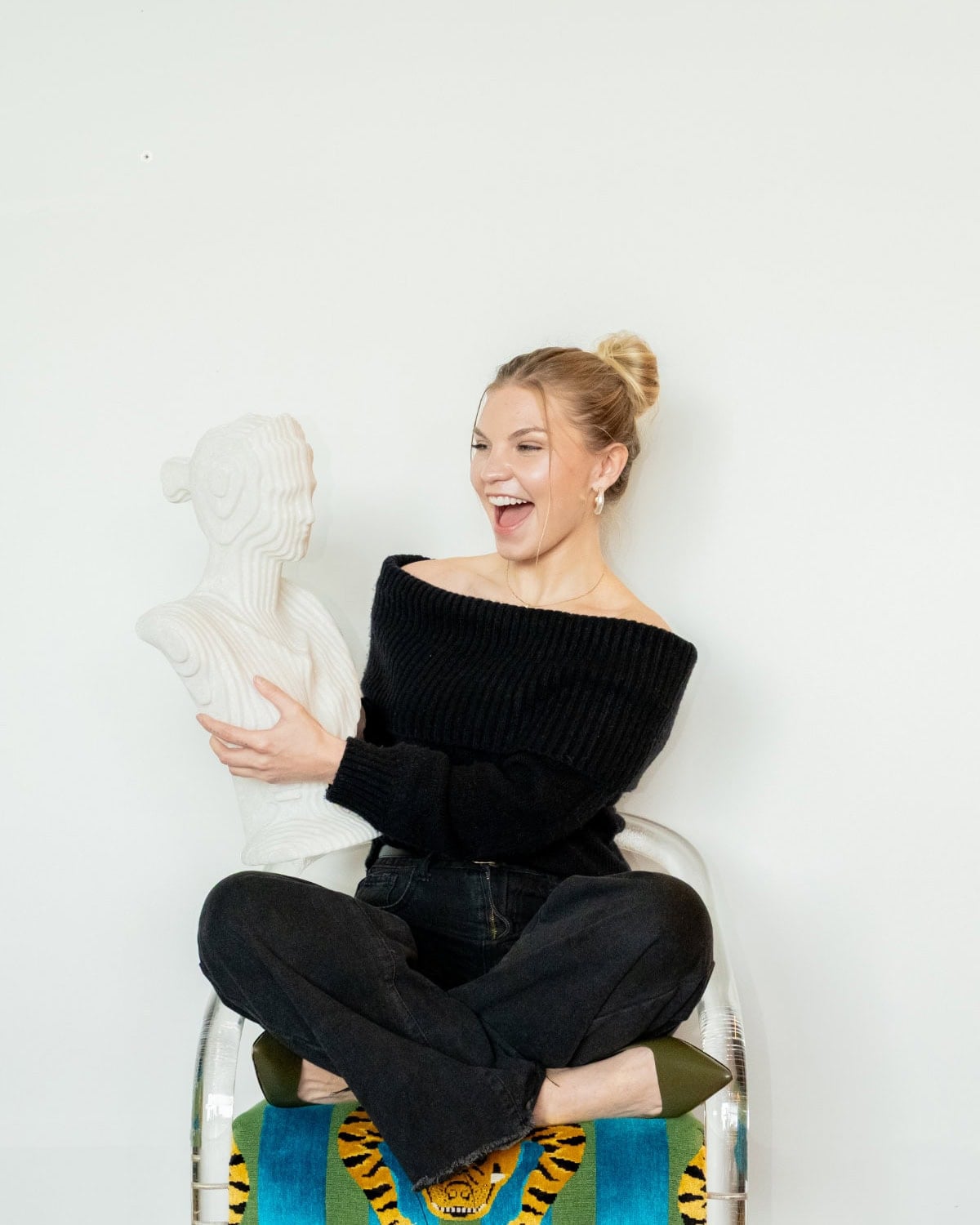 A woman sitting on a chair holds a white bust sculpture and smiles excitedly; there is a colorful fabric with a tiger design draped over the chair.