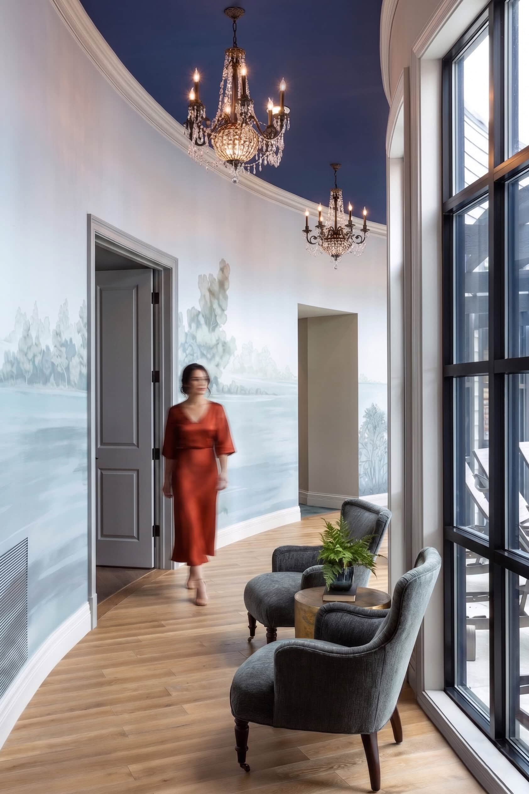 A woman in a red dress walks down a curved hallway with a mural wall, two chandeliers, and two gray armchairs by large windows.
