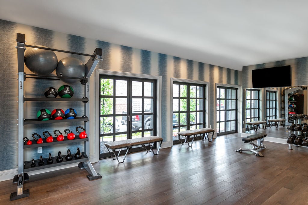 Modern gym with wooden floors, kettlebells, medicine balls, benches, large windows, and a wall-mounted TV.