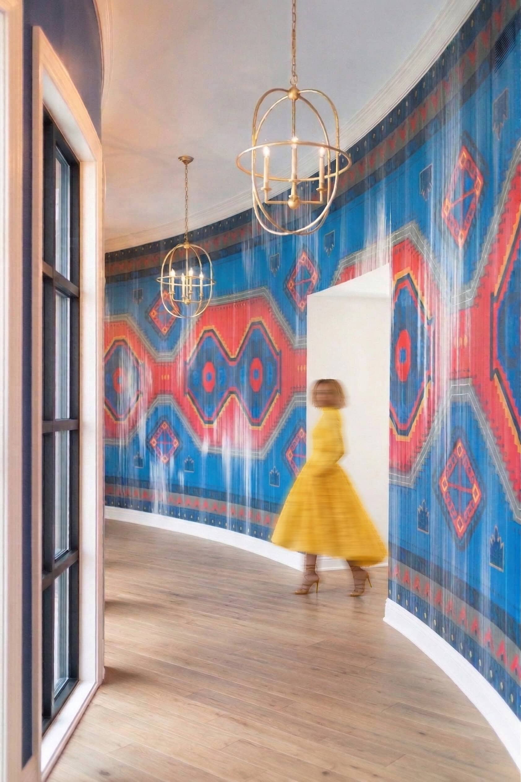 A person in a yellow dress walks down a curved hallway with wood floors, gold light fixtures, and blue and red geometric patterned wallpaper.