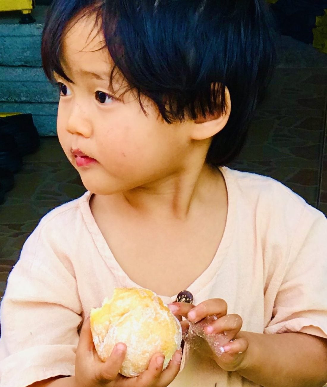 A young child with dark hair and a light shirt, styled by Crimson Design Group, holds a round pastry while looking to the side with a neutral expression.