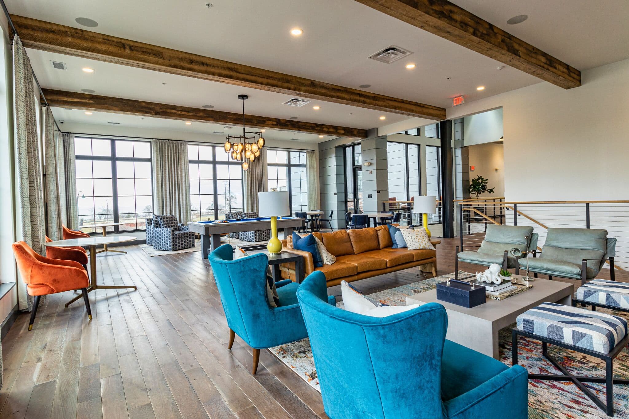 Spacious modern lounge with wooden floors, colorful chairs, couches, large windows, exposed beams, and decorative lighting.