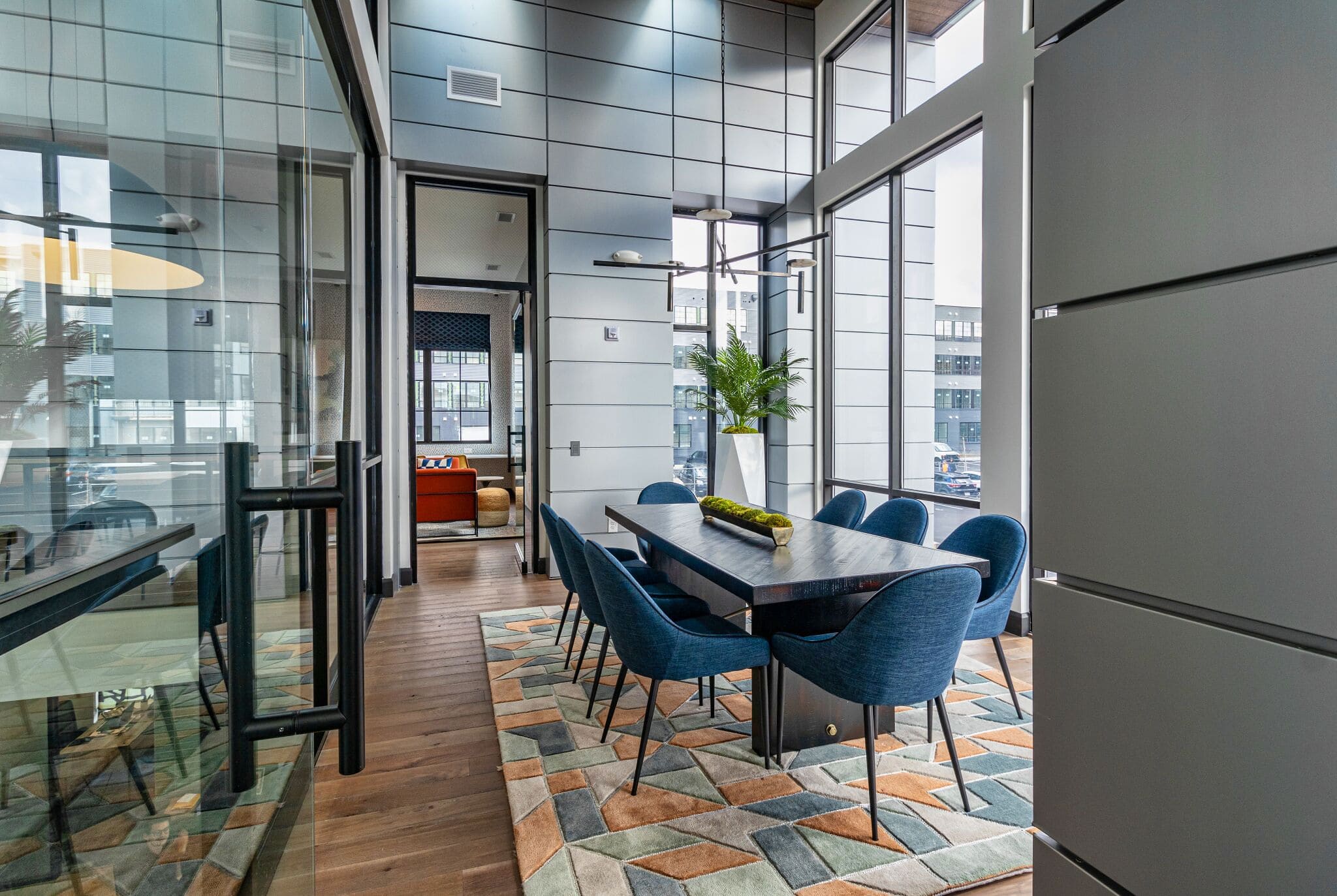 Modern conference room with large windows, blue chairs around a black table, geometric rug, and a plant centerpiece; adjacent office visible through glass wall.