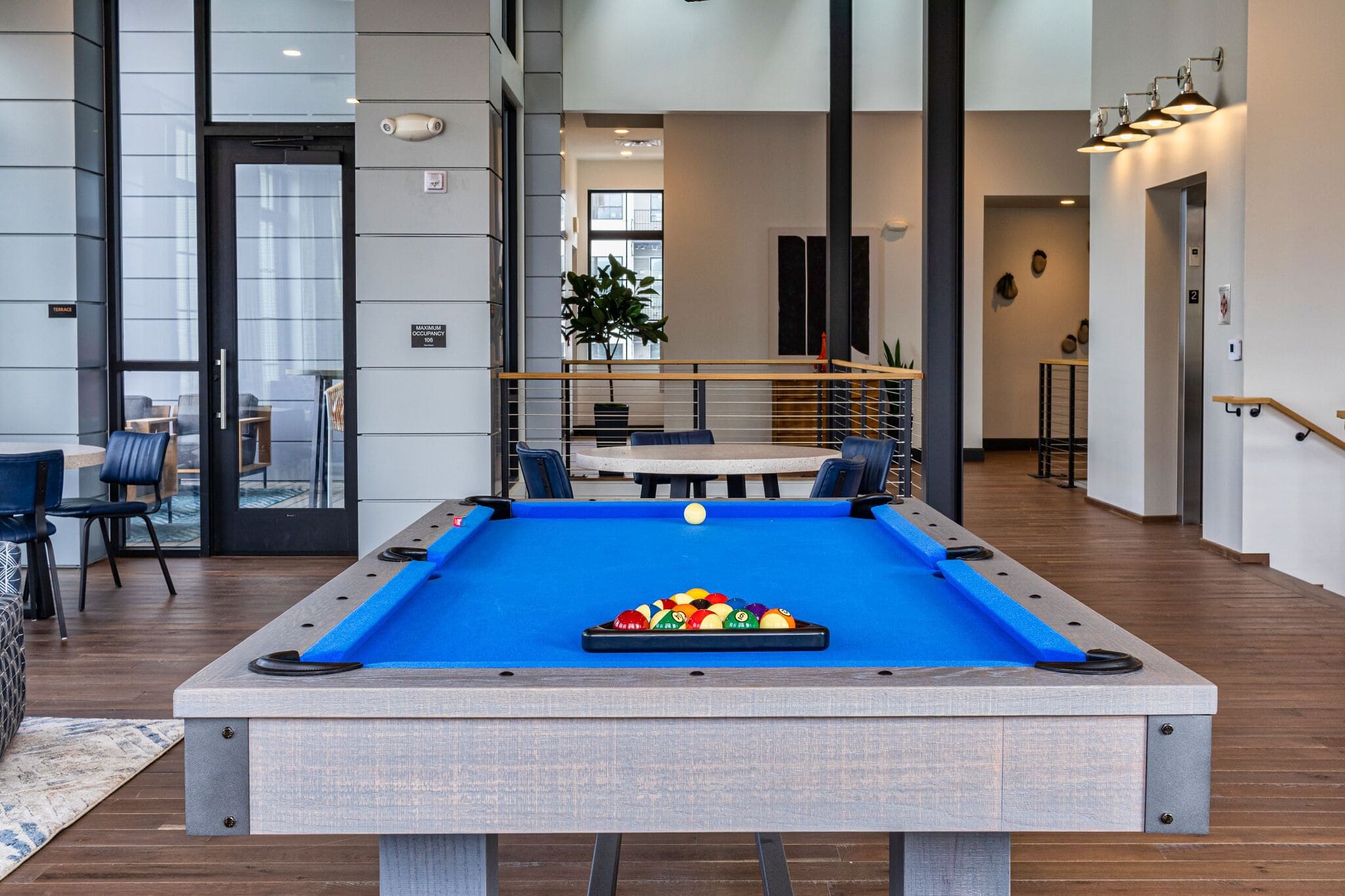 A modern indoor space featuring a blue pool table set up for a game, with chairs and tables in the background and large windows letting in natural light.