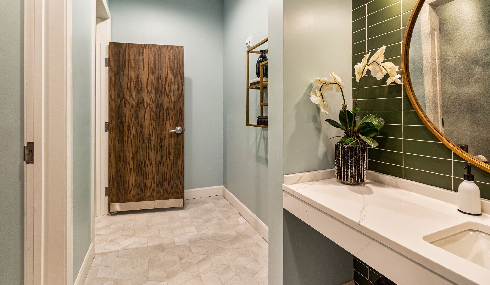 A modern bathroom with a wood door, hexagonal tile floor, a vanity with a white countertop, a round mirror, and a potted orchid.
