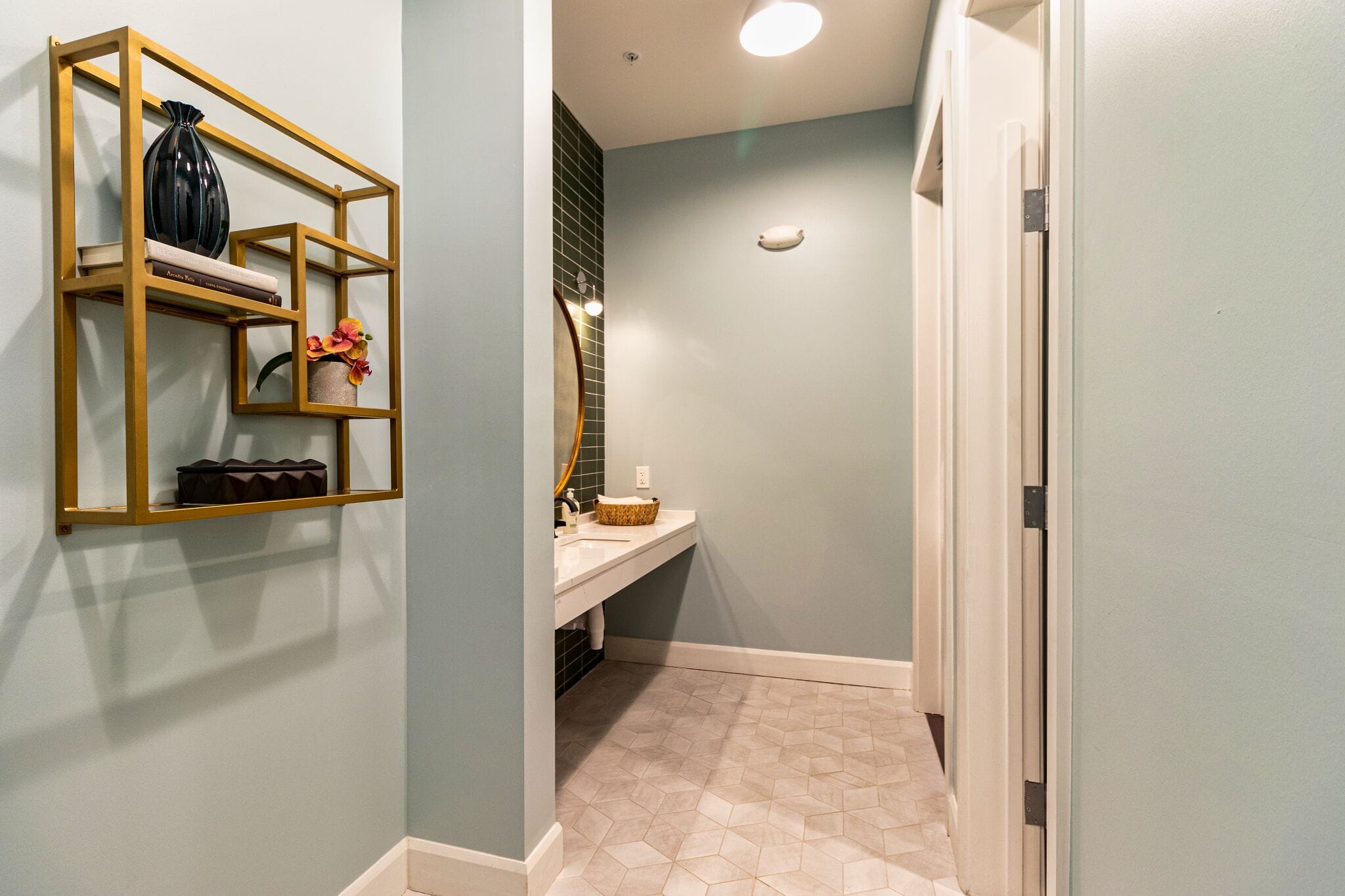 Modern bathroom with light blue walls, geometric floor tiles, a large round mirror above a white counter, and a gold wall shelf holding decor items.
