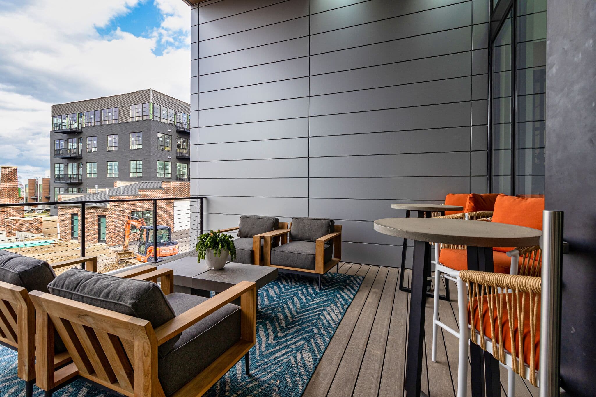 Modern outdoor balcony with wooden furniture, gray cushions, orange pillows, a potted plant, and a blue rug; urban buildings are visible in the background.