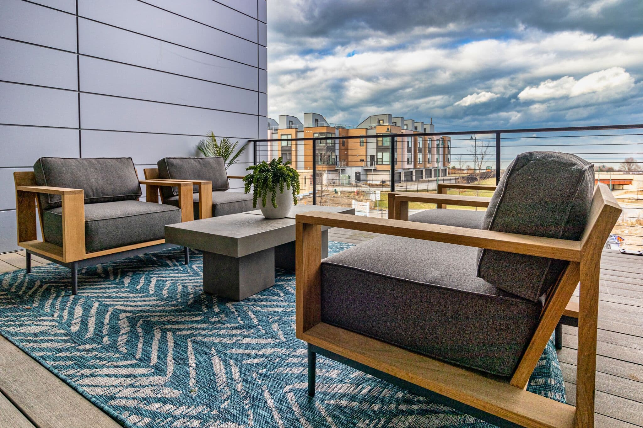 Modern outdoor patio with gray cushioned chairs, a concrete table, and a blue patterned rug, overlooking buildings and cloudy sky in the background.