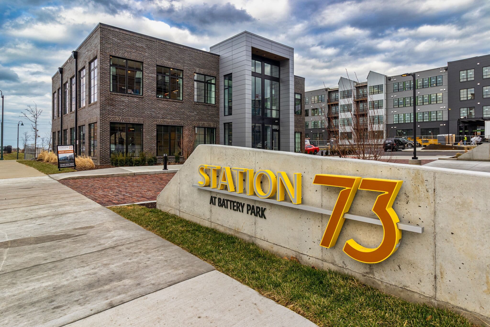 Modern commercial building with large sign reading "Station 73 at Battery Park" on a concrete wall; multi-story apartments visible in the background under a cloudy sky.
