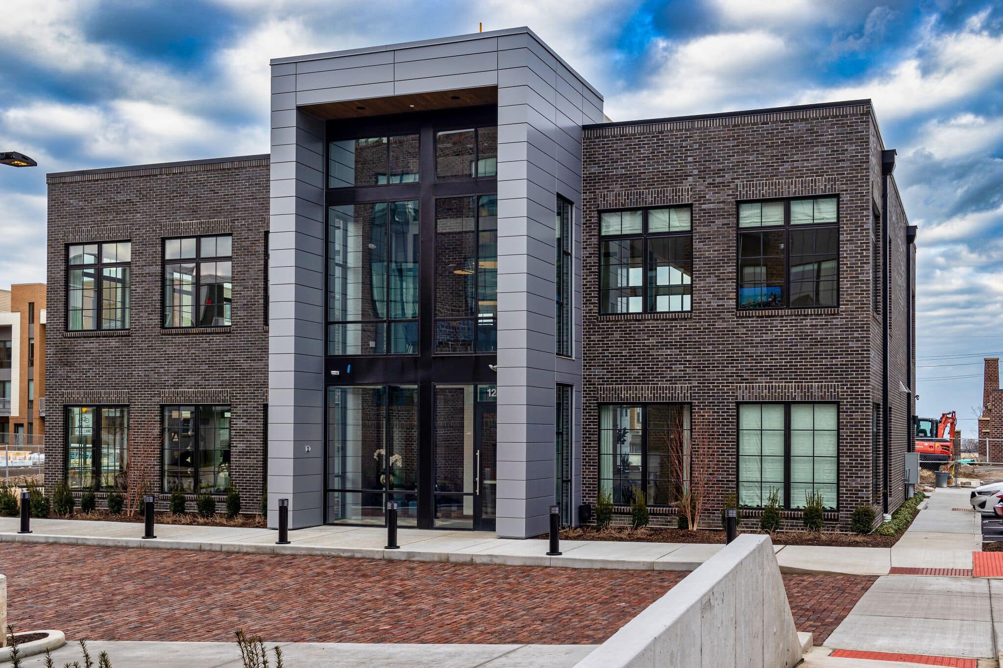 Two-story modern commercial building with dark brick exterior, large windows, and a central glass entrance, set on a paved street under a partly cloudy sky.