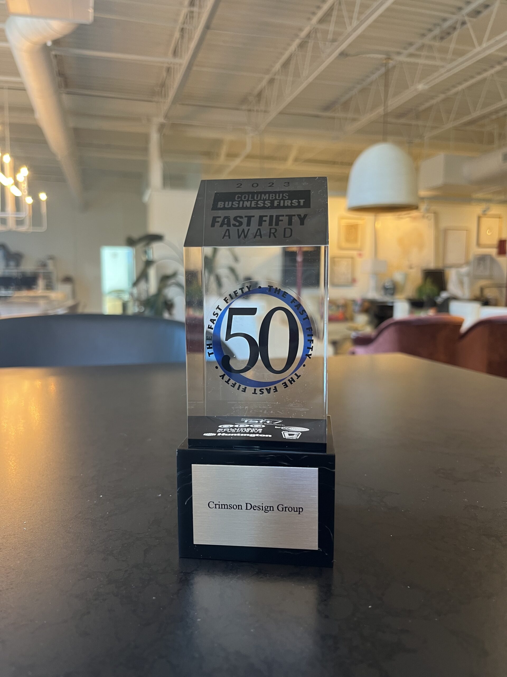A glass trophy labeled "Fast Fifty Award" sits on a table in an office, proudly displaying "Crimson Design Group" at its base, celebrating the company’s outstanding achievement.
