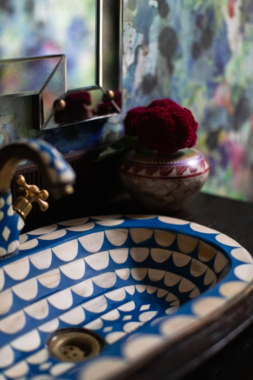 Blue and white patterned sink with brass faucet, small mirror, and a vase with dark red flowers on a counter against a colorful, abstract-patterned wall.